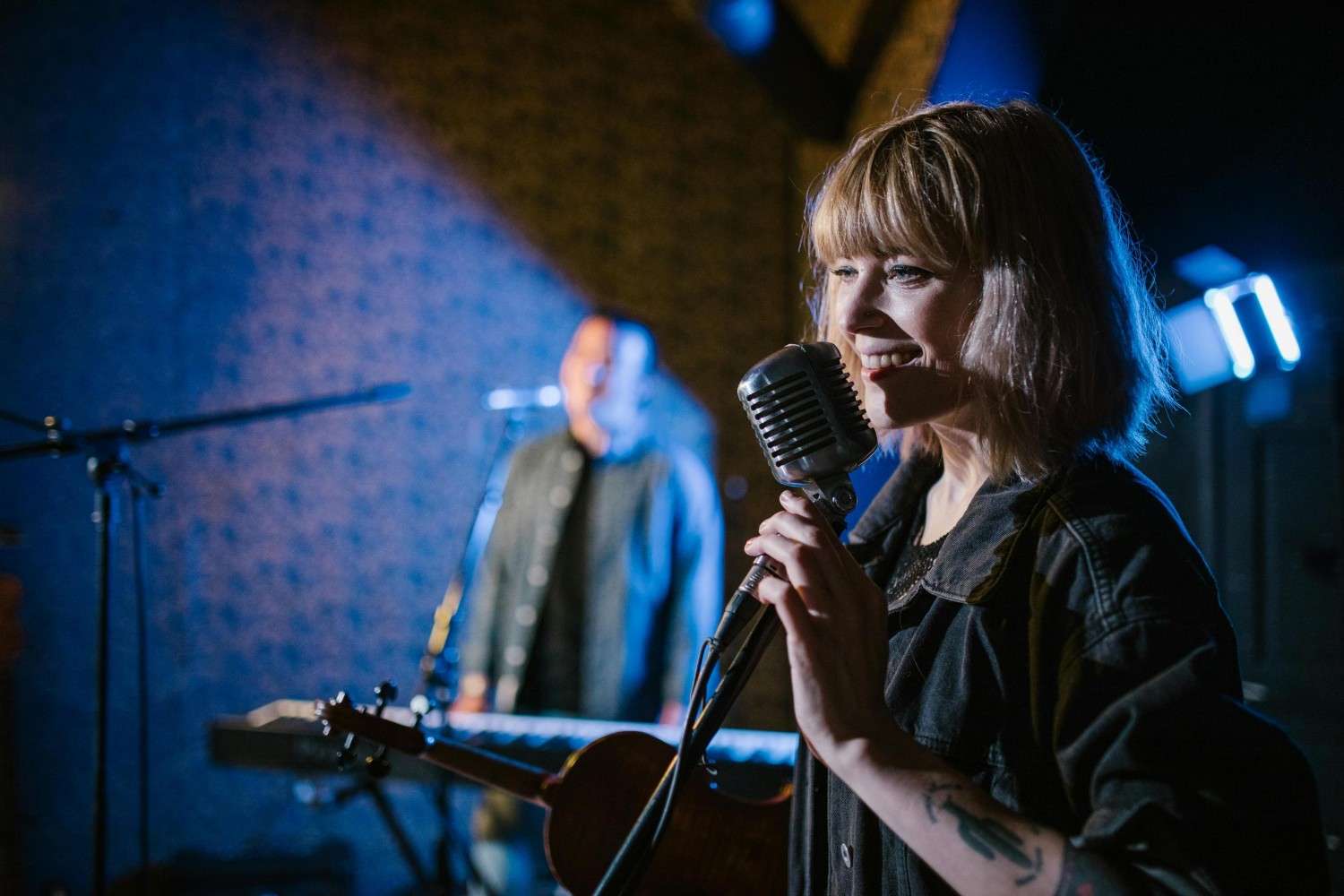 A smiling vocalist with a blonde bob sings into a vintage silver microphone while holding a violin, illuminated by soft stage lighting. In the background, a fellow musician stands at a keyboard bathed in blue light, contributing to the atmospheric live performance setting.