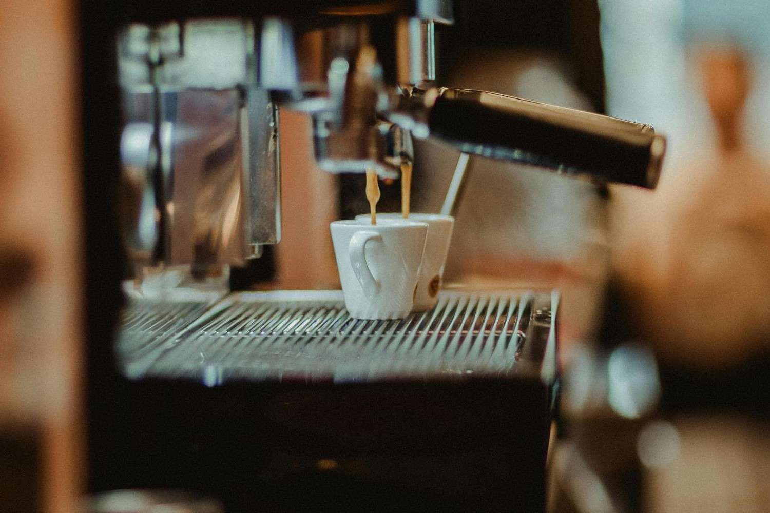 A close-up captures an espresso machine in action, pouring steady streams of hot coffee into two small white cups resting on a metal grate. The shallow depth of field softens the background, highlighting the gleaming stainless steel and the rich color of the brew.