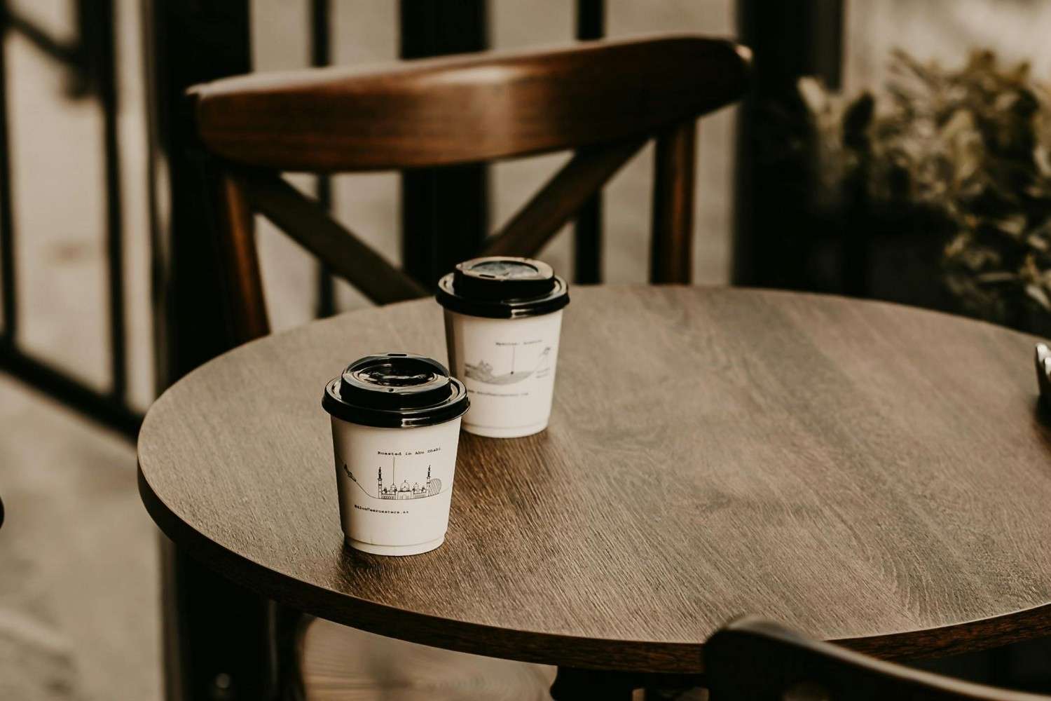 Two white paper cups with black lids, featuring minimalist architectural illustrations, sit on a round, dark wooden table. A wooden chair and blurred background elements frame the scene, evoking a quiet and warm café atmosphere.