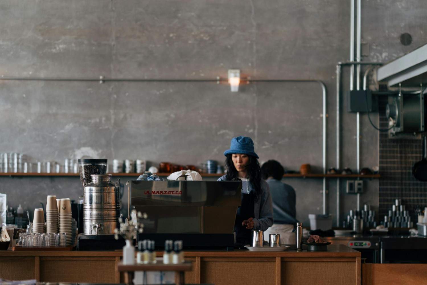 A barista wearing a blue bucket hat stands behind a wooden counter, focused on her tasks near a large espresso machine. The industrial-style café features textured grey walls and exposed piping, with neat stacks of paper cups and glassware lining the workspace.