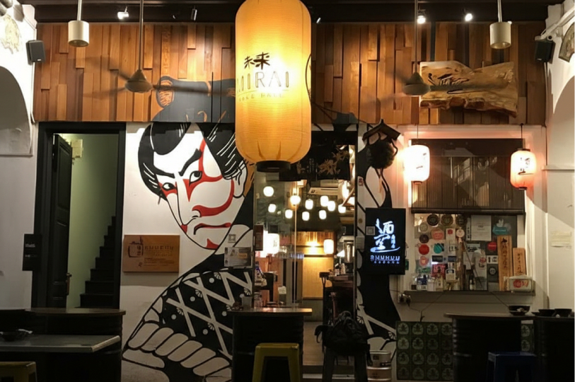 An outdoor seating area of a Japanese izakaya featuring a large, stylized wall mural of a Kabuki actor, glowing yellow and white paper lanterns, and wood-paneled accents above the entrance.