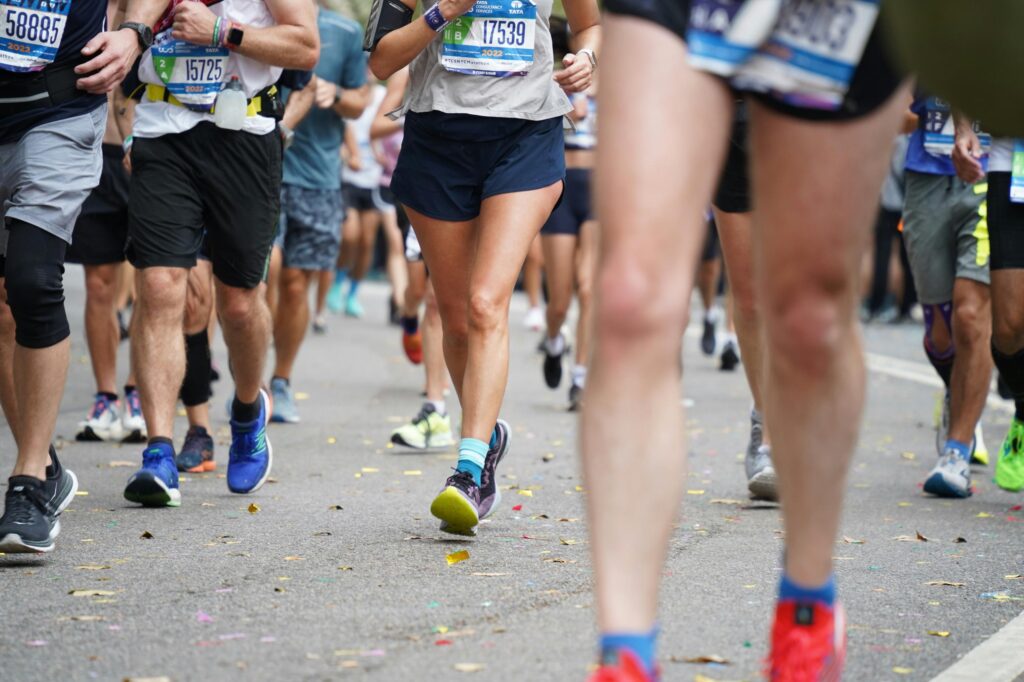 This eye-level, close-up shot captures a crowded pack of runners competing in a marathon on an asphalt road scattered with colorful confetti. The focus remains on the lower bodies and legs of the participants, highlighting various styles of athletic gear and colorful running shoes as they move forward in unison.