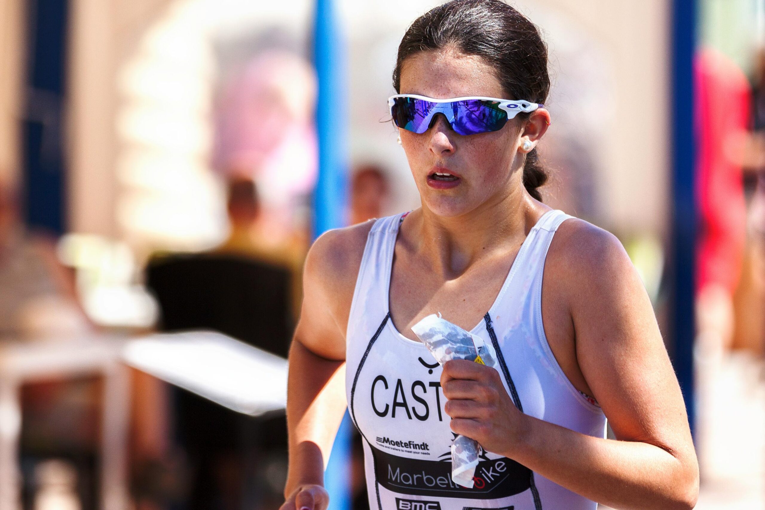 This medium shot features a female athlete in a white racing singlet and blue-tinted sunglasses, captured in mid-stride during a race. She holds a small, crumpled packet in her hand while maintaining a focused expression against a brightly lit, blurred outdoor background.