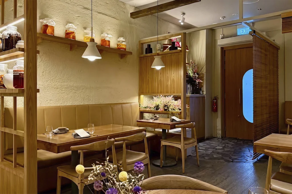An inviting, warm-toned restaurant interior featuring light wood furniture and textured cream walls. A long tan leather banquette runs along the wall beneath wooden floating shelves lined with large glass jars of amber-colored infusions. Two minimalist white pendant lights hang over the wooden tables. To the right, a wooden door with a large circular blue-tinted window is partially obscured by a bamboo roll-up screen, while a bouquet of dried purple and yellow flowers sits in the blurred foreground.