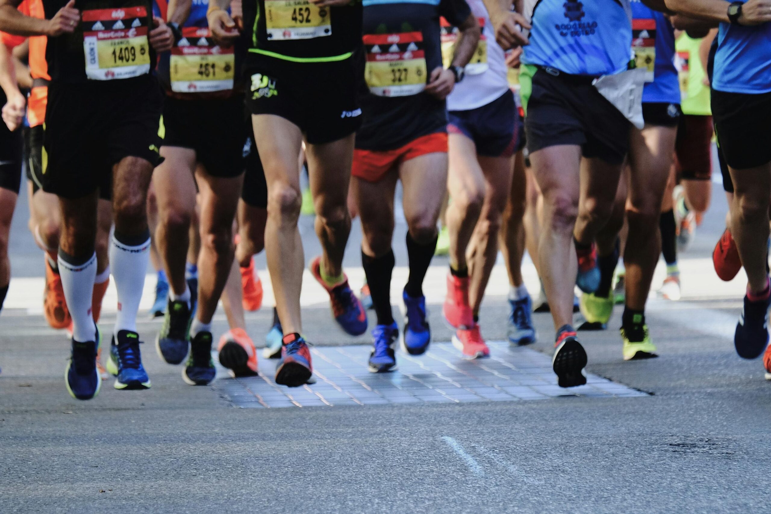  This low-angle, close-up shot focuses on the legs and feet of a group of marathon runners as they cross a specialized timing mat on an asphalt road. The image captures the dynamic movement of various colorful running shoes and athletic gear, emphasizing the intensity and synchronized pace of the race participants.