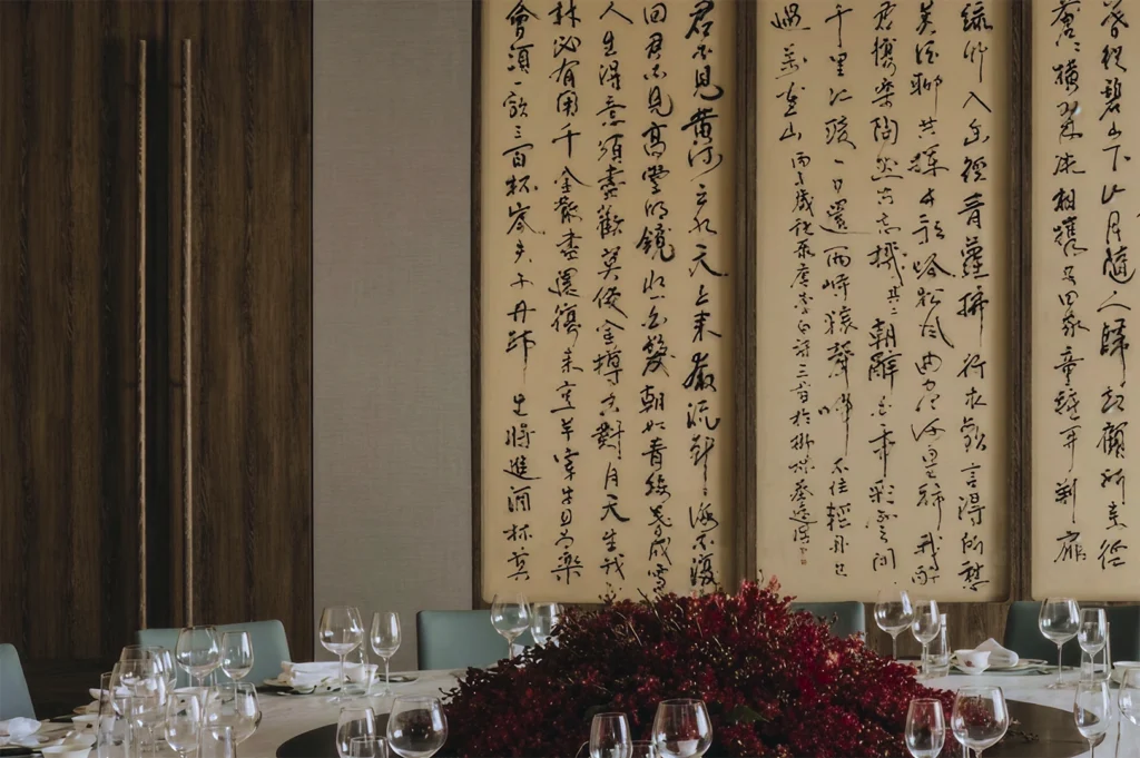 A close-up shot of an elegant, large round dining table set for a formal meal. The center of the table is dominated by a massive, lush arrangement of deep red textured flowers. In the background, three tall, vertical hanging scrolls feature intricate black ink Chinese calligraphy on aged cream paper. The room is framed by dark wood paneling and features light turquoise upholstered chairs, with numerous polished wine and water glasses catching the soft, ambient light.