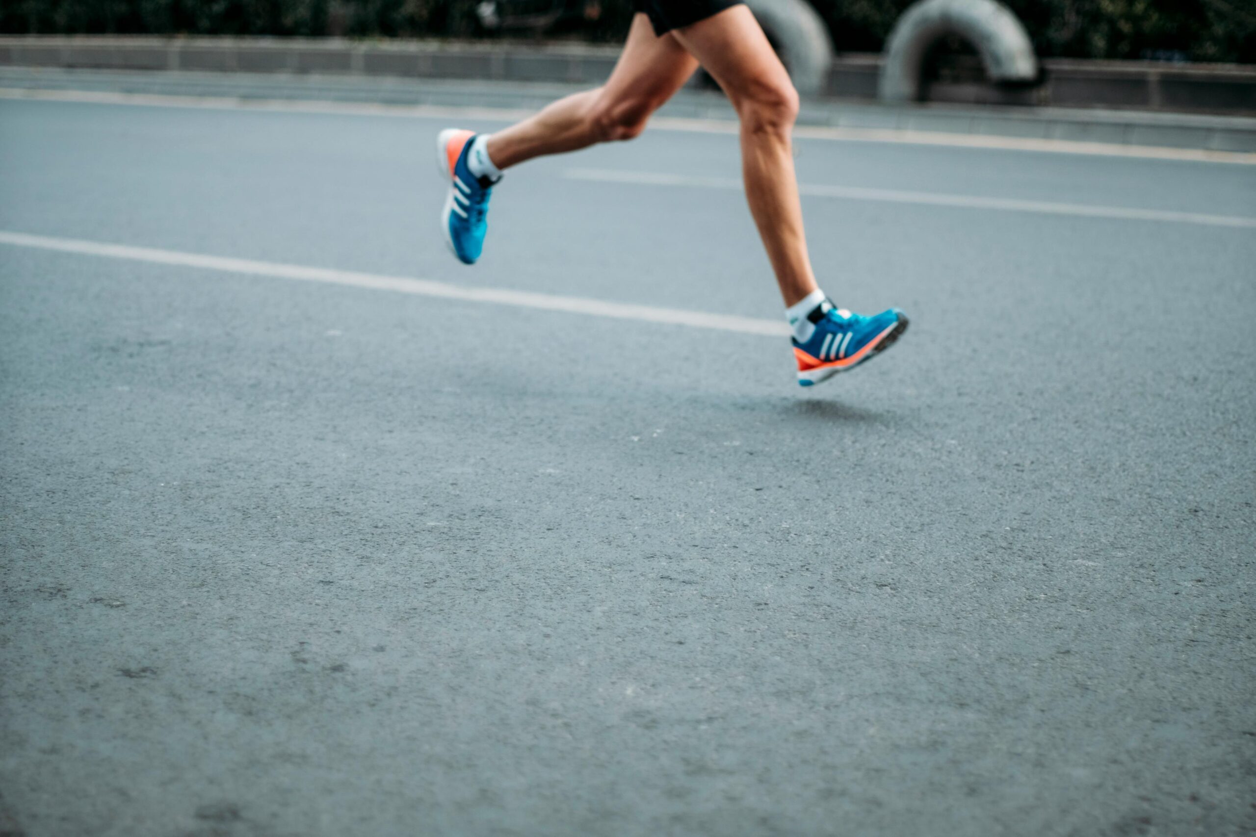  This eye-level, side-view shot captures the legs of a runner in mid-stride on a gray asphalt road. The athlete is wearing bright blue running shoes with white and orange accents, emphasizing a sense of speed and athletic motion against the blurred background.