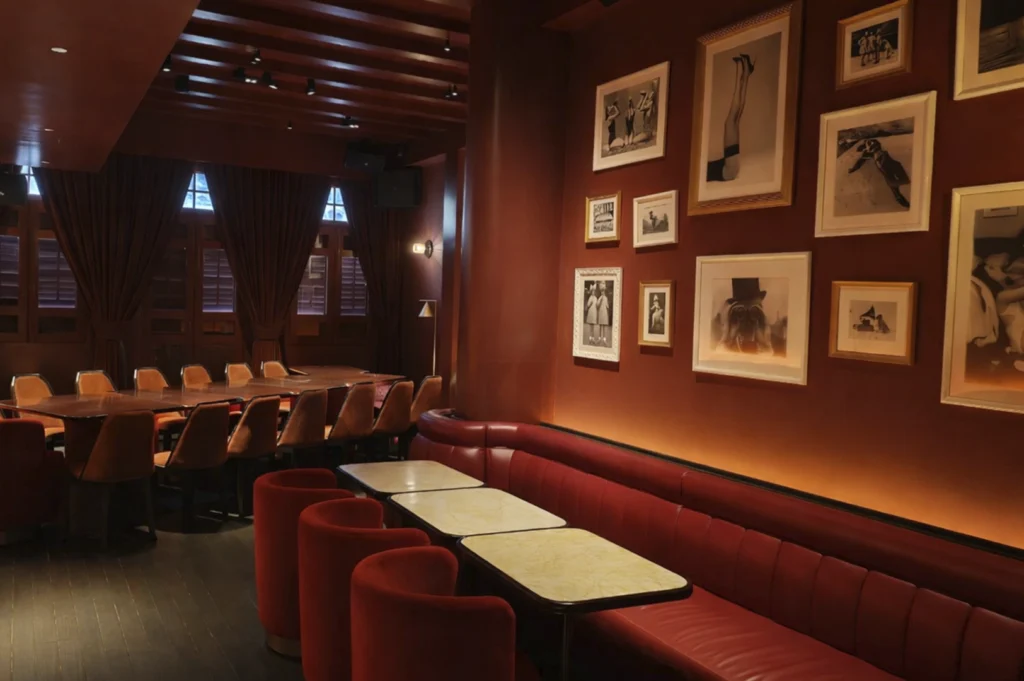 A moody, sophisticated lounge or private dining room bathed in warm red light. The left side of the room features a long dark wooden table with orange velvet chairs. On the right, a plush red leather banquette follows the curve of a deep terracotta-colored wall, which is adorned with an organized gallery of black-and-white art photography in various white and gold frames. Three small marble-topped cafe tables with red velvet stools are lined up in front of the banquette under a dark, beamed wooden ceiling.