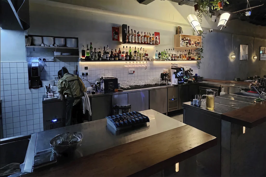 A behind-the-scenes view of a modern professional bar or open kitchen area. The space features industrial stainless steel workstations and dark wood countertops. In the background, a staff member in a green uniform is busy at a prep station near white-tiled walls. Above the back counter, several floating wooden shelves are stocked with a wide variety of liquor bottles, illuminated by warm under-shelf lighting. The ceiling is dark and exposed, featuring hanging greenery and unique, angled white pendant lamps.