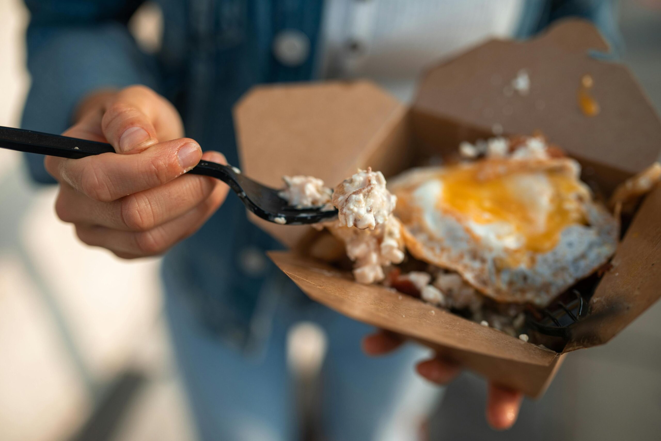 A person wearing a denim jacket holds an open cardboard takeout box containing a hearty meal of fries and cheese topped with a fried egg. One hand grips a black plastic fork, lifting a bite of the food towards the viewer while the background remains softly blurred.