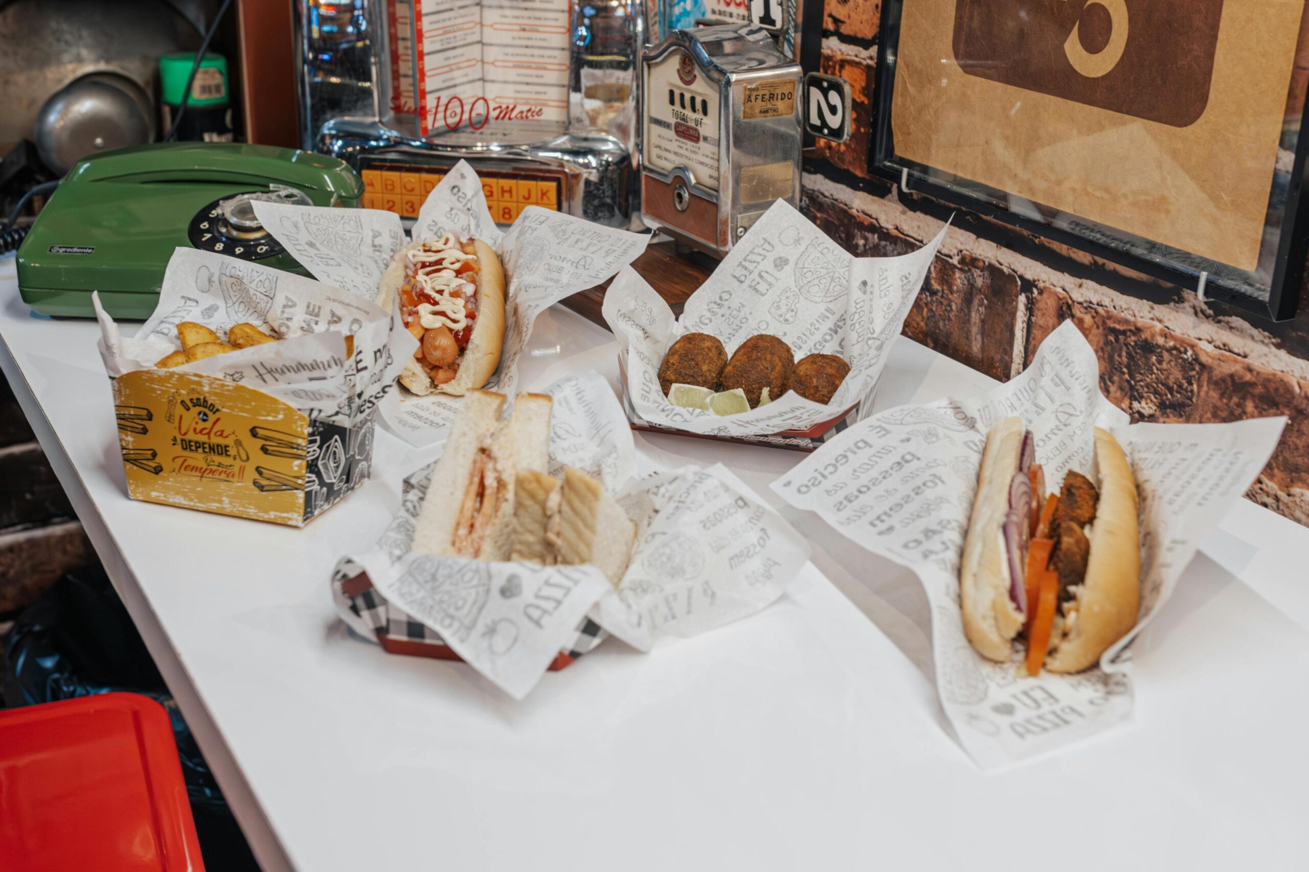 A spread of fast food items, including hot dogs, sandwiches, french fries, and croquettes, is arranged on paper wrappers across a white countertop. The background evokes a retro diner atmosphere, featuring an exposed brick wall decorated with a green rotary telephone and a miniature vintage jukebox.