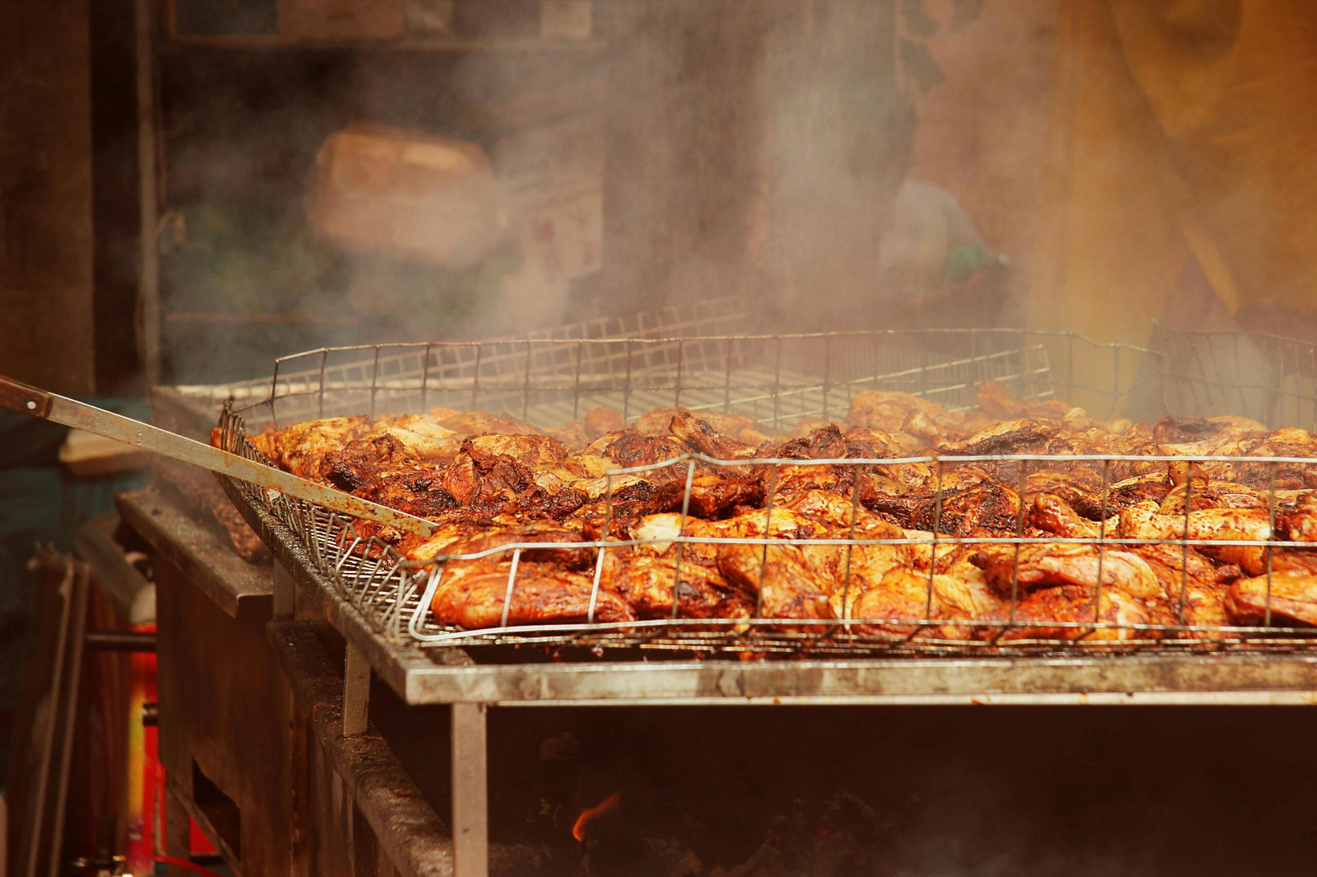 Thick smoke rises from a large metal grate filled with seasoned chicken pieces roasting over an open fire. A long metal tool reaches in from the left to tend the sizzling meat, which is browned and charred from the intense heat.