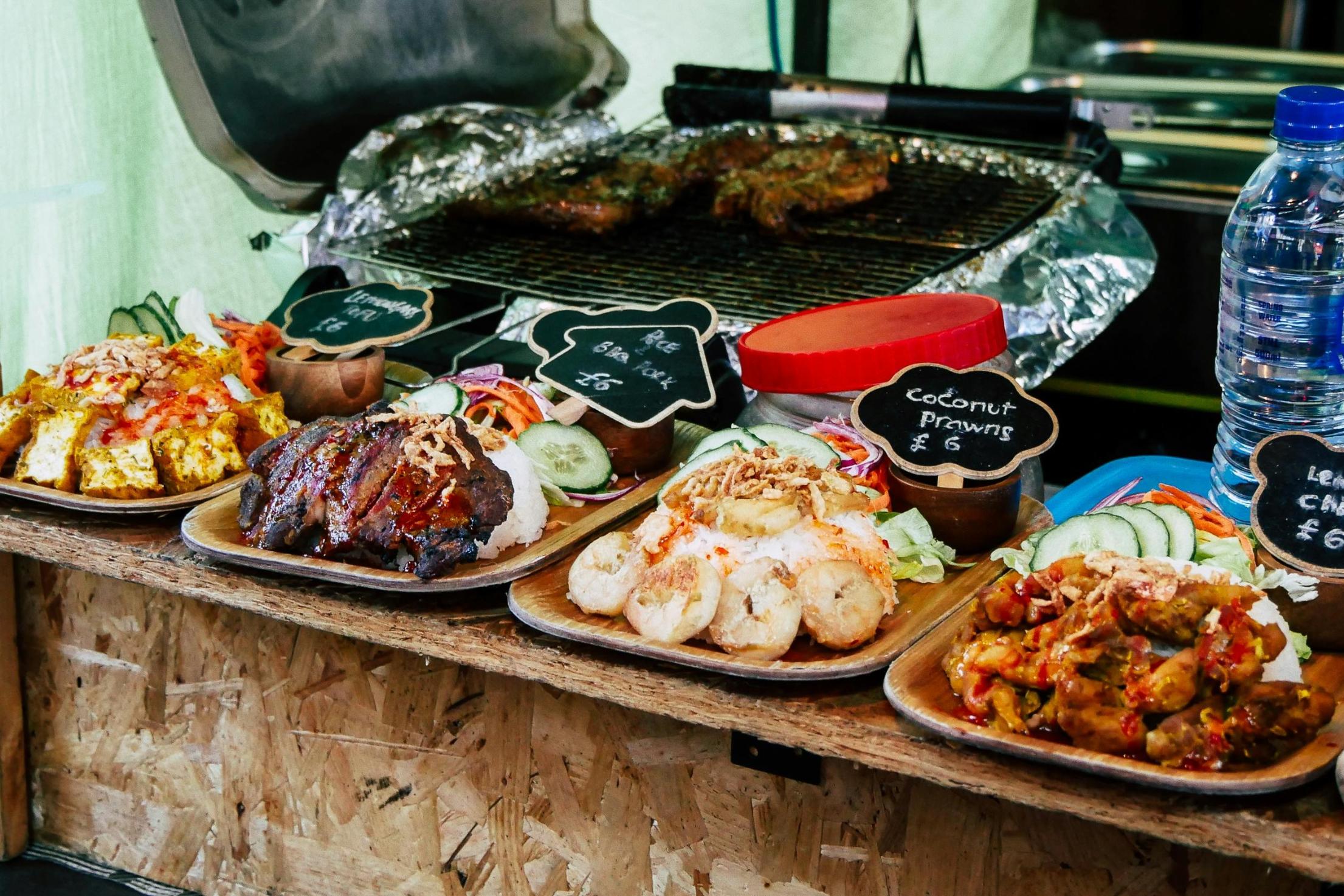 A selection of street food dishes, such as coconut prawns and BBQ pork served with rice, lines a wooden counter, each accompanied by a small blackboard sign indicating the price. In the background, pieces of meat roast on a metal grill rack, while a large plastic water bottle stands near the food display.