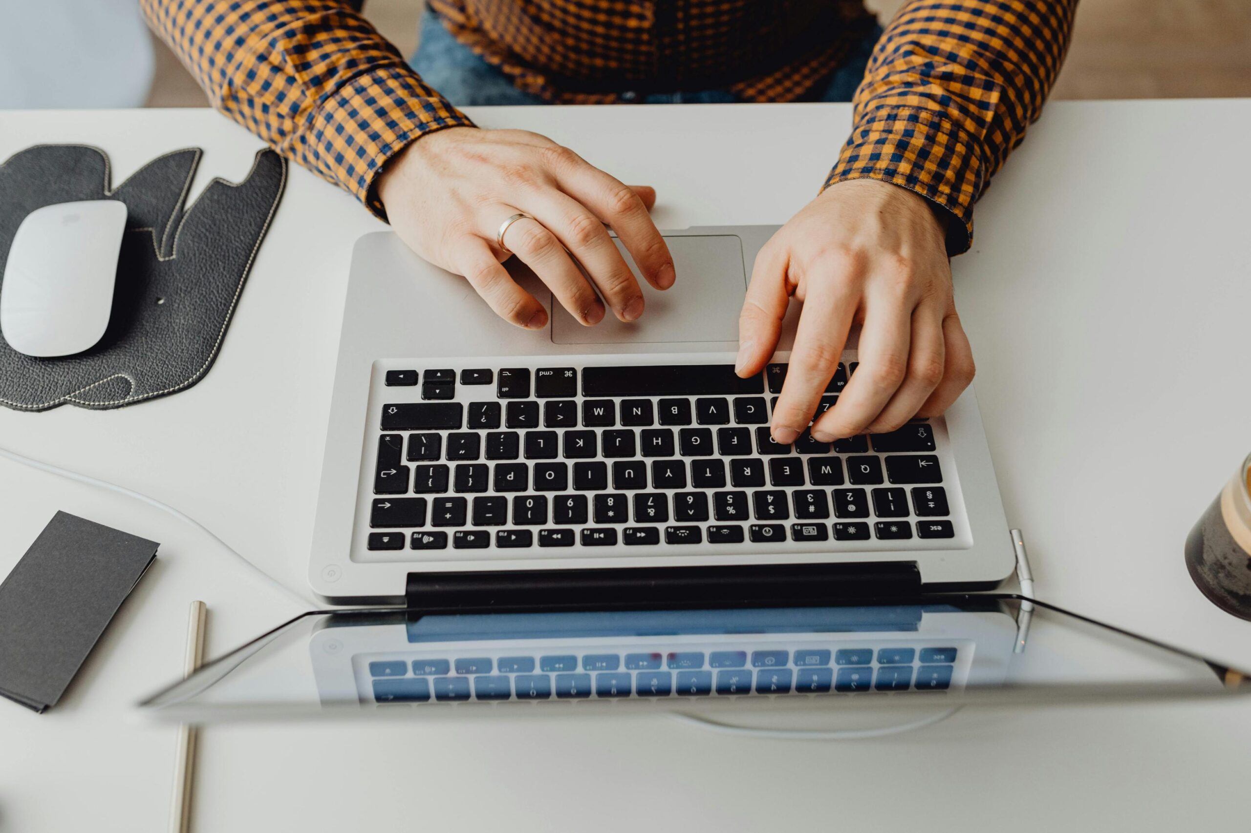 Hands of a man in a yellow plaid shirt typing on a laptop keyboard at a white office desk.