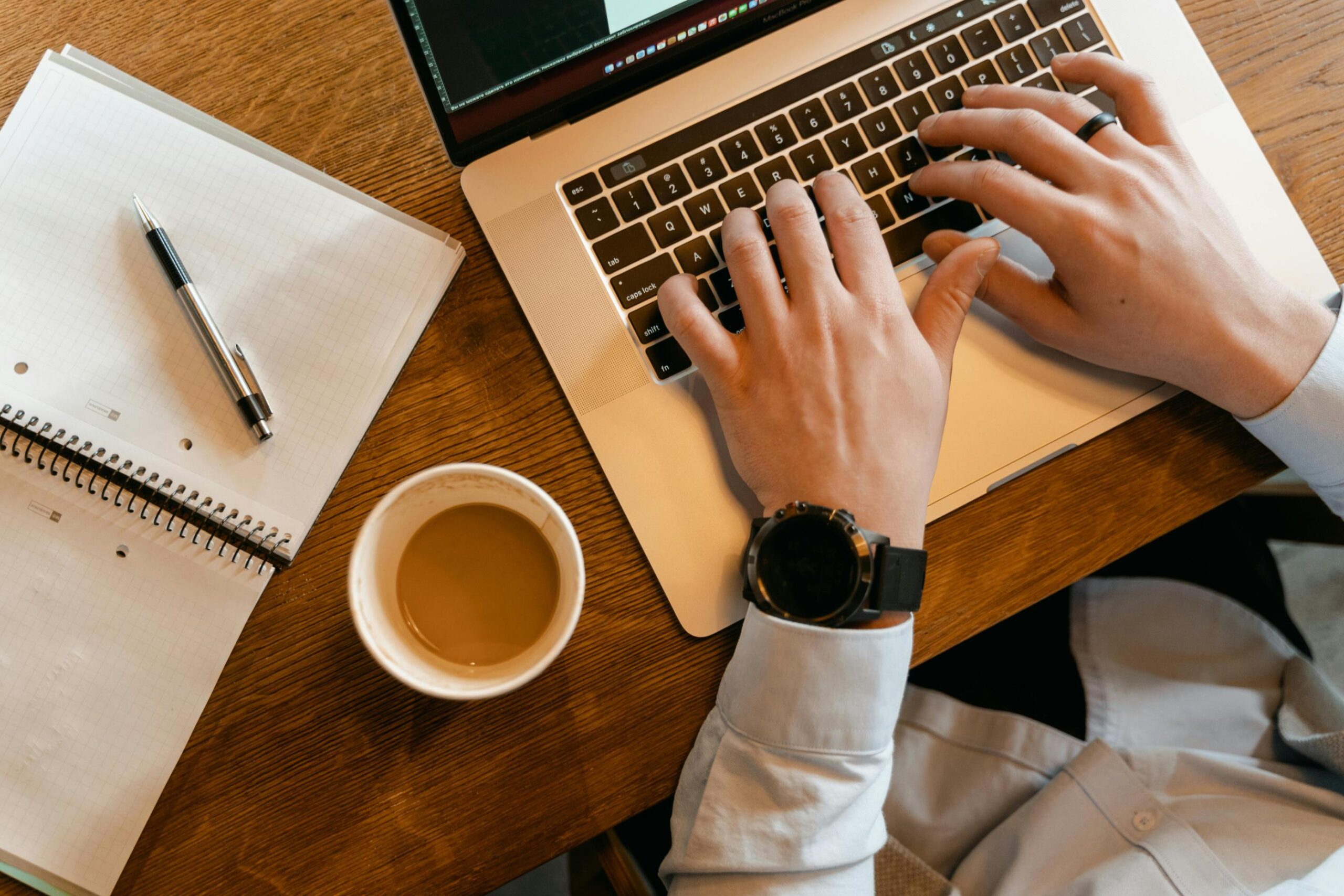 Overhead view of a man typing on a laptop at a wooden desk with a notebook and coffee.