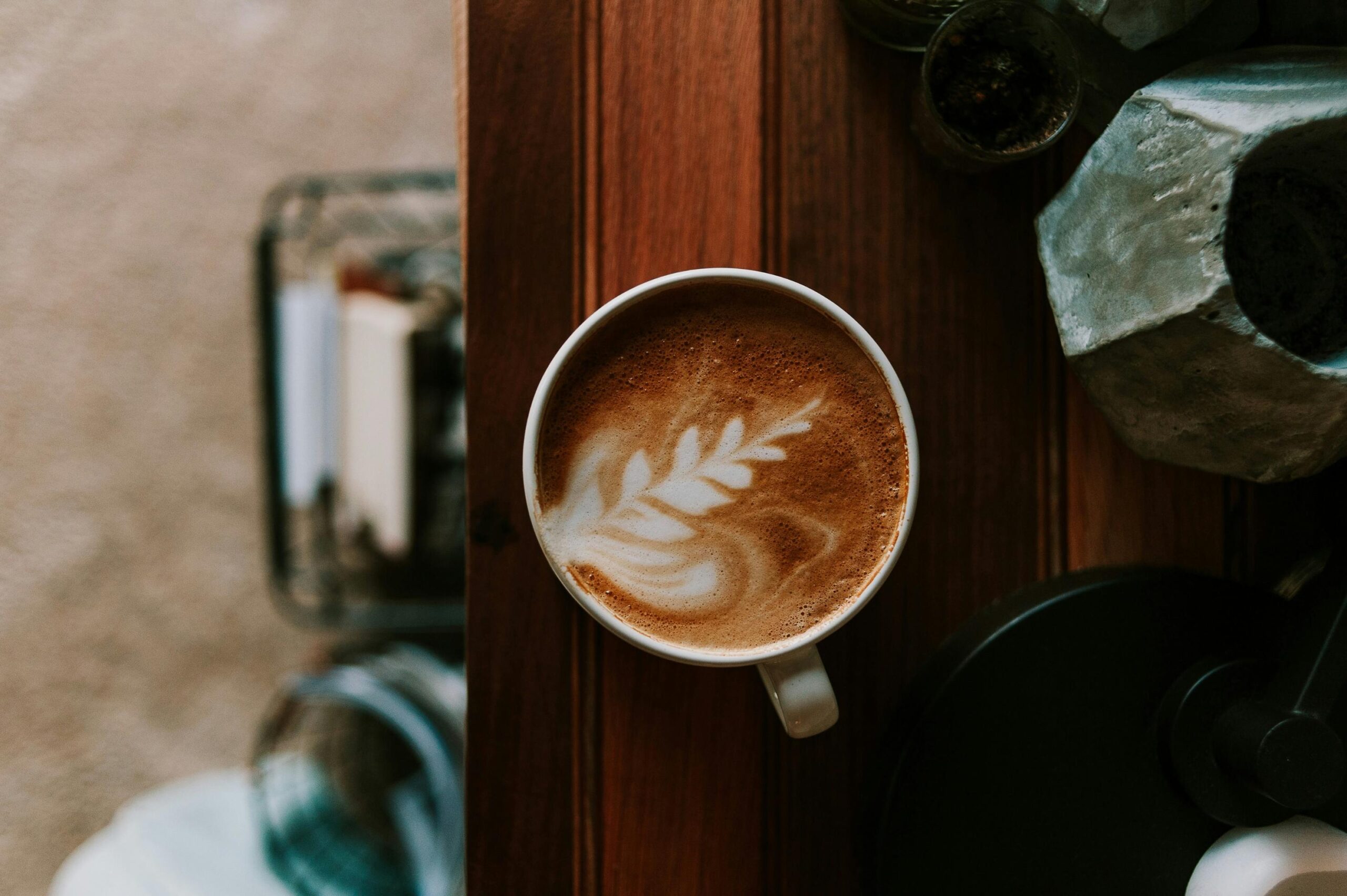 A white ceramic cup filled with a latte featuring a delicate fern design sits near the edge of a dark wooden table. Next to the coffee, a geometric concrete planter adds texture to the scene, while the floor below remains softly out of focus.