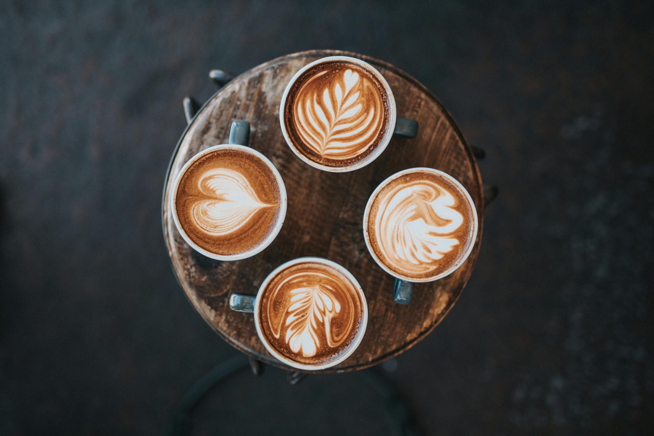 Four ceramic cups of coffee are arranged in a tight circle on a small, distressed wooden stool, each showcasing a unique style of latte art. The designs range from delicate leaves to a swan and a heart, standing out brightly against the rich brown espresso and the moody, dark background.