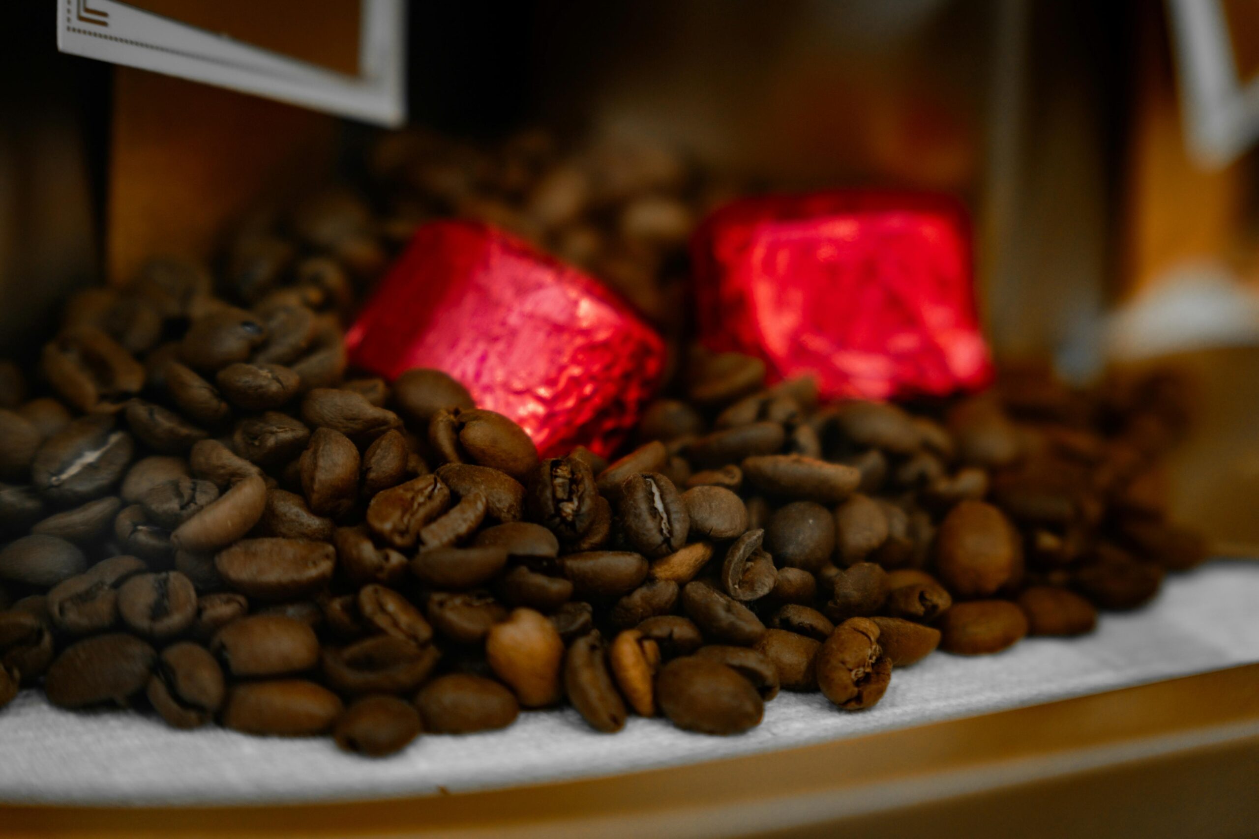 A collection of roasted coffee beans is piled high, creating a textured surface of warm brown tones. Two bright red, foil-wrapped candies rest atop the beans, contrasting sharply with the dark coffee surroundings.