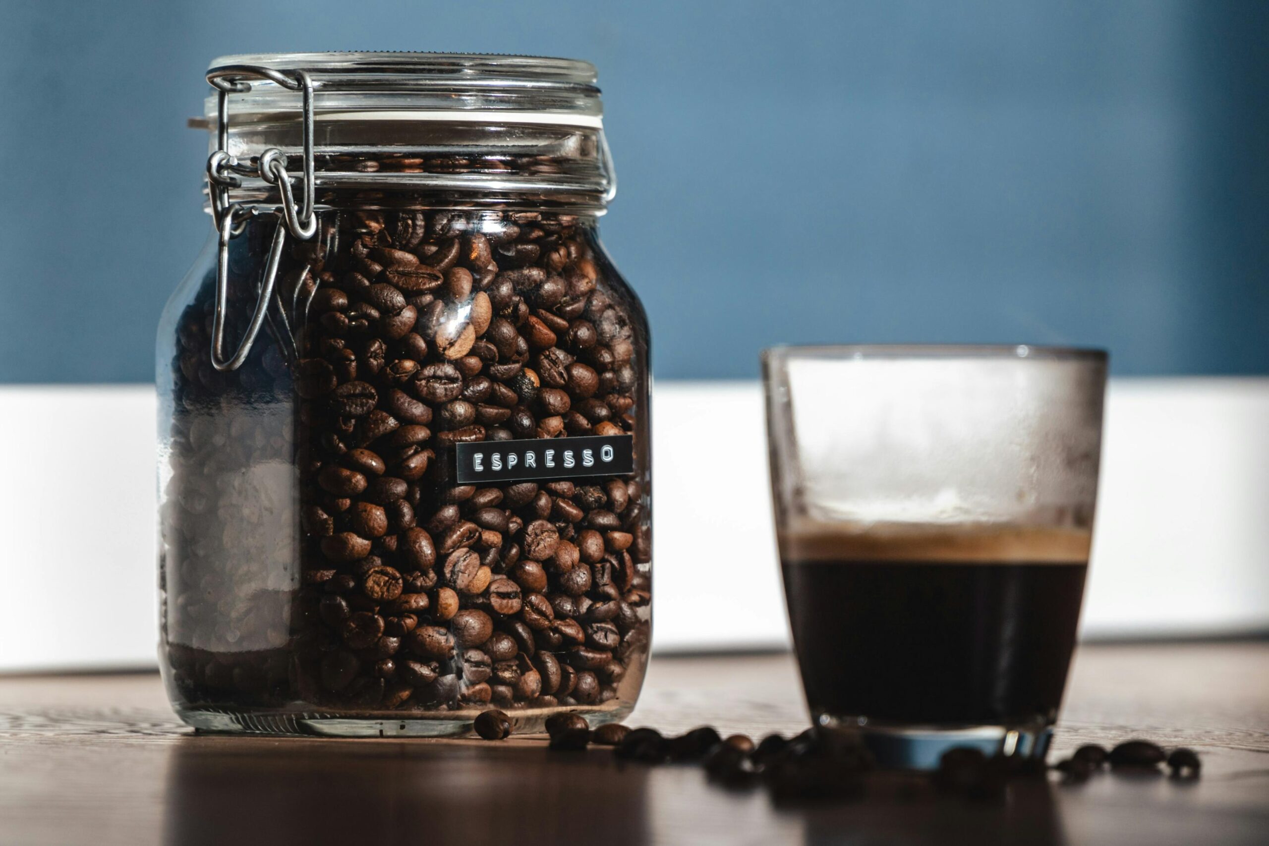 A glass jar filled with dark roasted coffee beans, labeled "ESPRESSO," sits next to a fresh cup of coffee on a wooden surface. Scattered beans surround the base of the jar and glass, contrasting against the blue and white background wall.