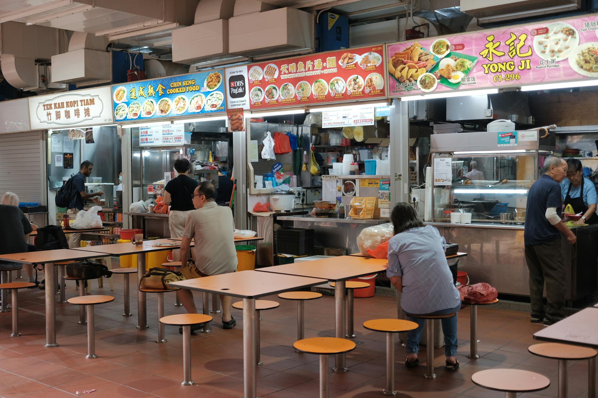 This image captures the casual atmosphere of a hawker center, featuring a row of food stalls adorned with bright, illustrated menu boards. Customers are seated at rows of simple tables with round stools, eating and interacting while vendors prepare food in the background.