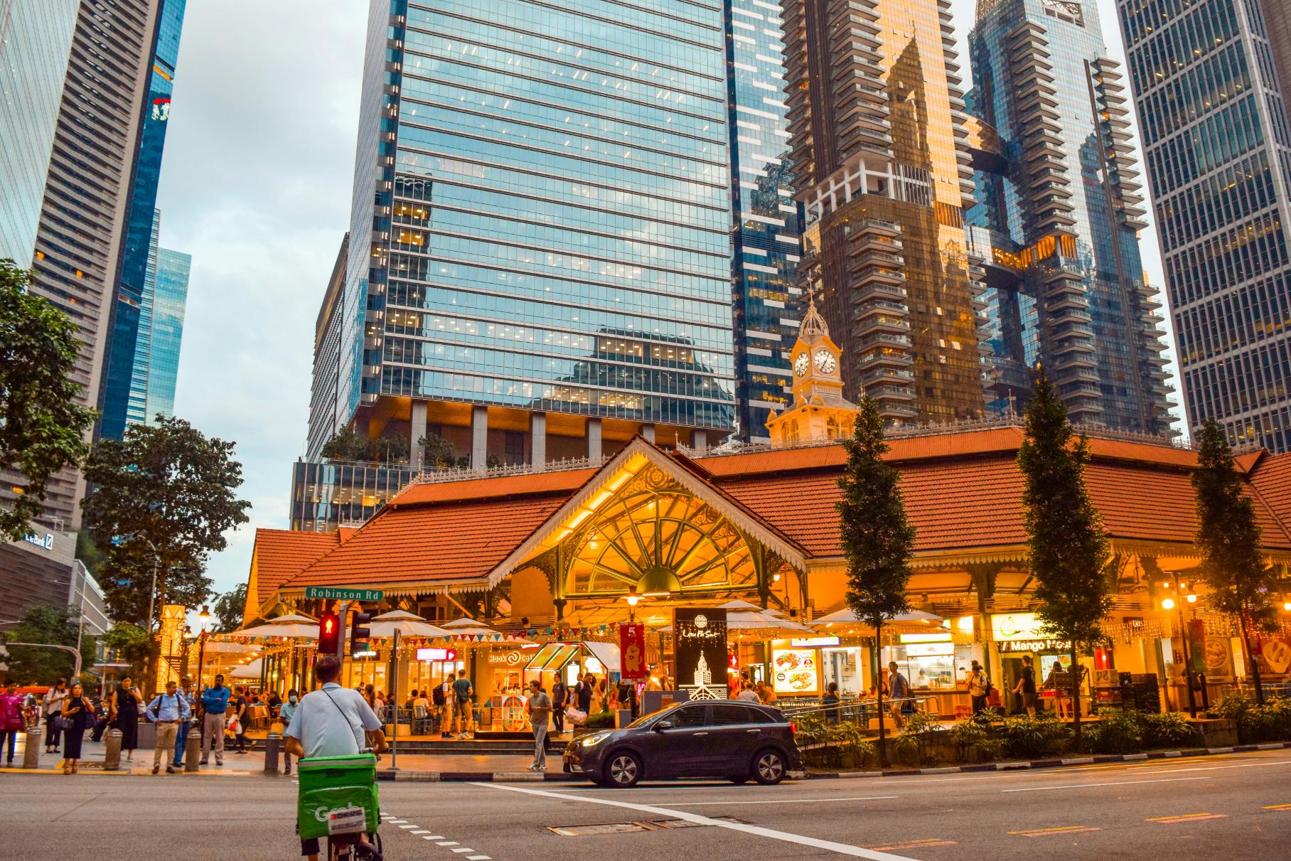 This image highlights a dramatic architectural contrast, featuring a historic, warm-lit market pavilion with an ornate clock tower nestled at the foot of towering modern skyscrapers. The scene captures a bustling evening atmosphere with pedestrians and traffic moving around the glowing heritage building set against the cool backdrop of the city skyline.