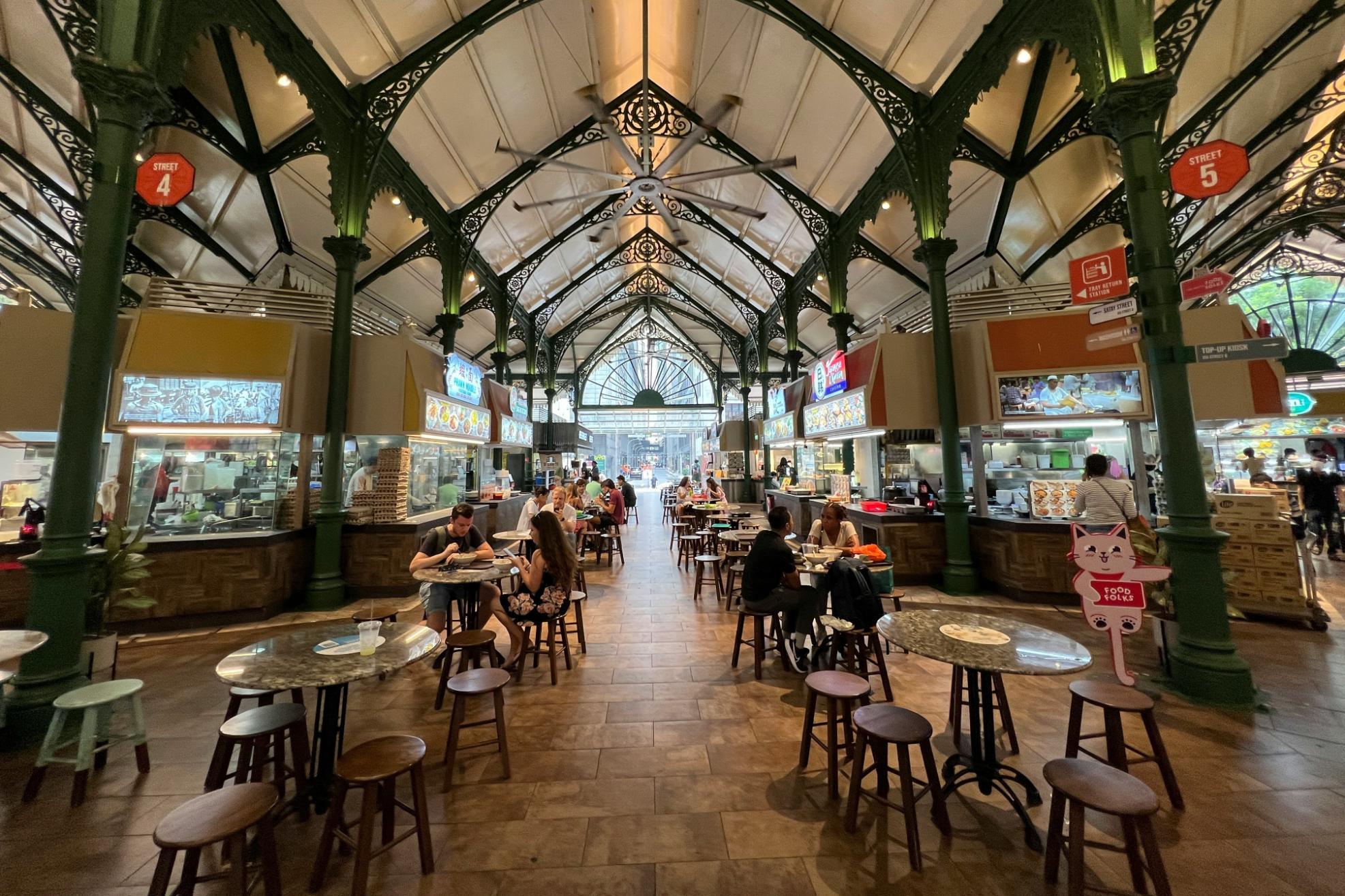 This image depicts the interior of a spacious, historic food hall featuring high vaulted ceilings supported by intricate green cast-iron columns and arches. Diners are seated at round tables along a wide central aisle, enjoying meals from the various food stalls lining the perimeter of the open-air structure.