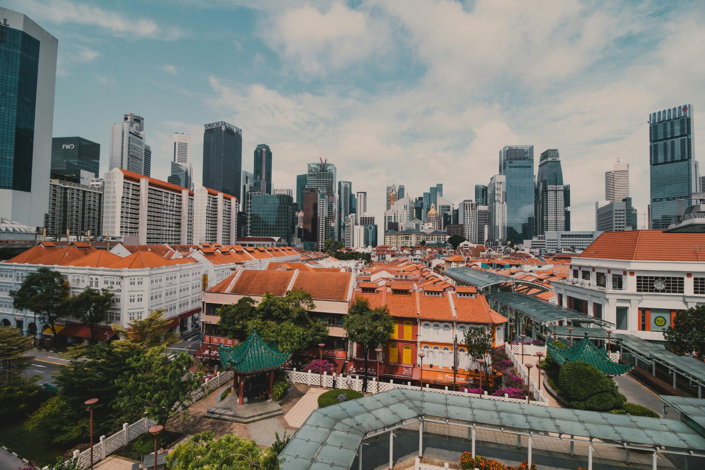 This cityscape image presents a striking juxtaposition of eras, with traditional low-rise buildings featuring orange terracotta roofs and ornate green pavilions dominating the foreground. In the background, a dense cluster of towering modern skyscrapers rises against the sky, creating a dramatic visual contrast between the city's heritage and its contemporary financial district.