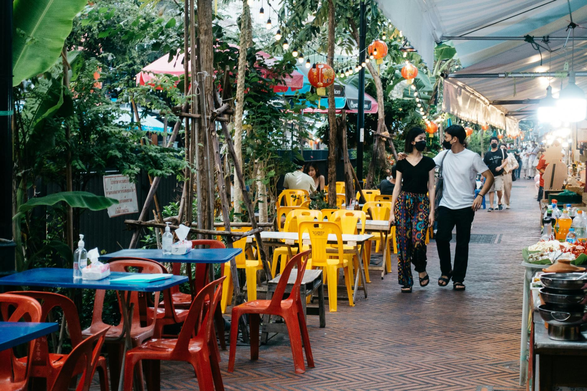 This image depicts a charming outdoor dining promenade at dusk, where vibrant red and yellow plastic chairs are nestled amidst lush greenery and glowing string lights. A couple walks arm-in-arm along the brick-paved path, enjoying the relaxed atmosphere of the market lined with food stalls.