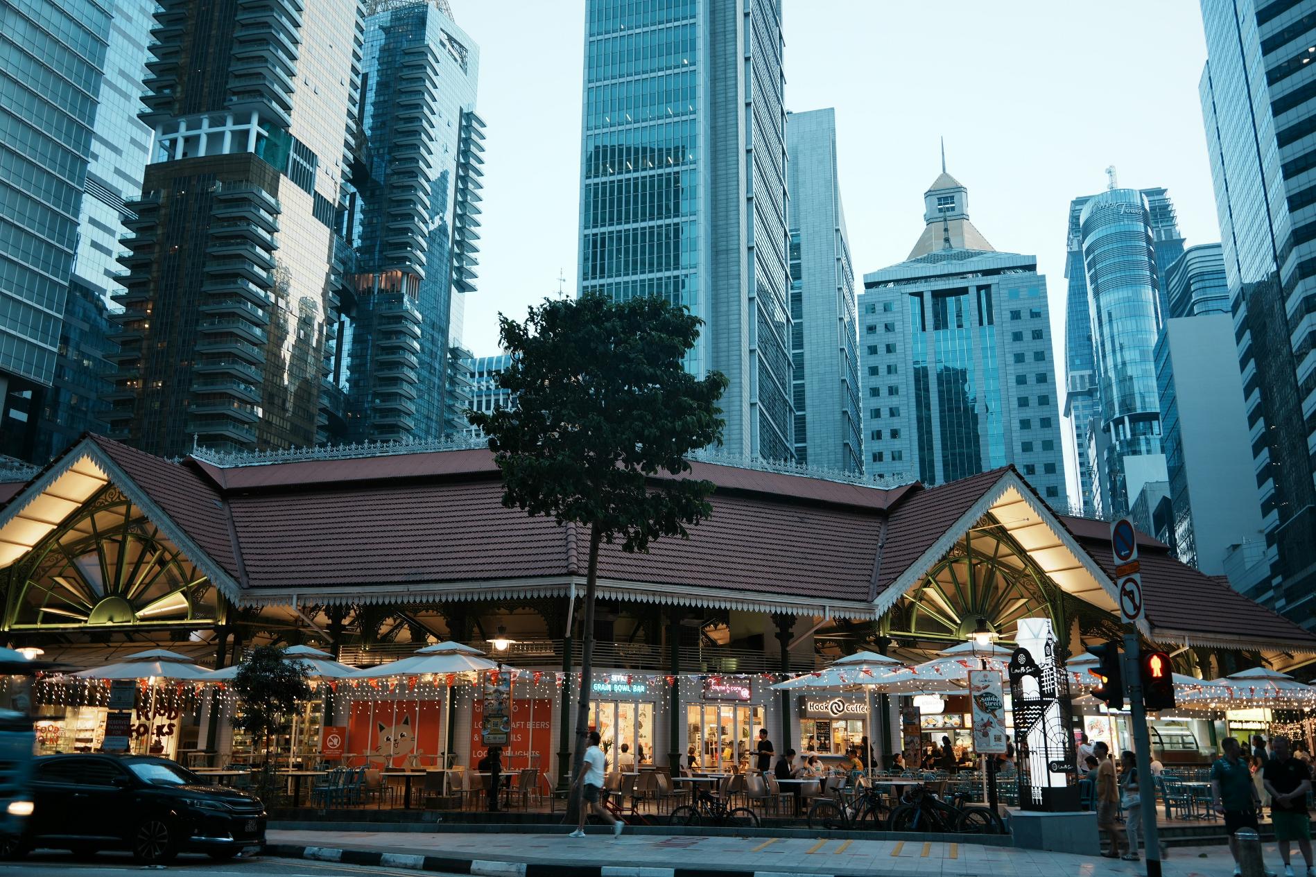 This urban landscape showcases a dramatic contrast between eras, featuring a historic, low-rise market building with a distinctive red tiled roof surrounded by soaring glass skyscrapers. Warm lights glow from the bustling open-air dining area at street level, creating a cozy atmosphere that stands out against the cool, blue tones of the modern city architecture above.