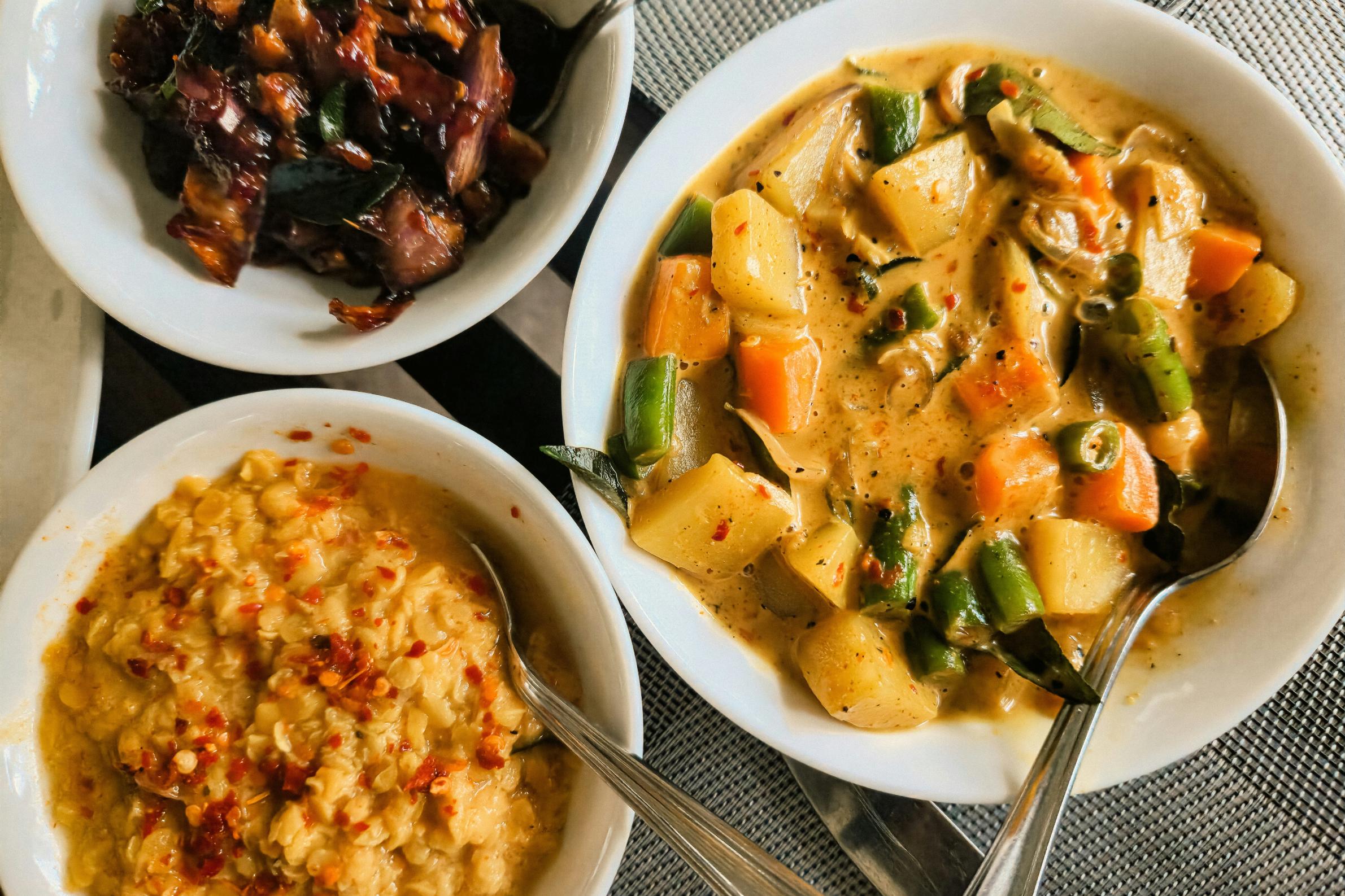 This overhead view presents a trio of white bowls containing a hearty meal, featuring a creamy vegetable curry loaded with potatoes, carrots, and green beans. The spread is complemented by a bowl of yellow lentils garnished with red chili flakes and a side of dark, savory fried vegetables, all set upon a textured gray surface.