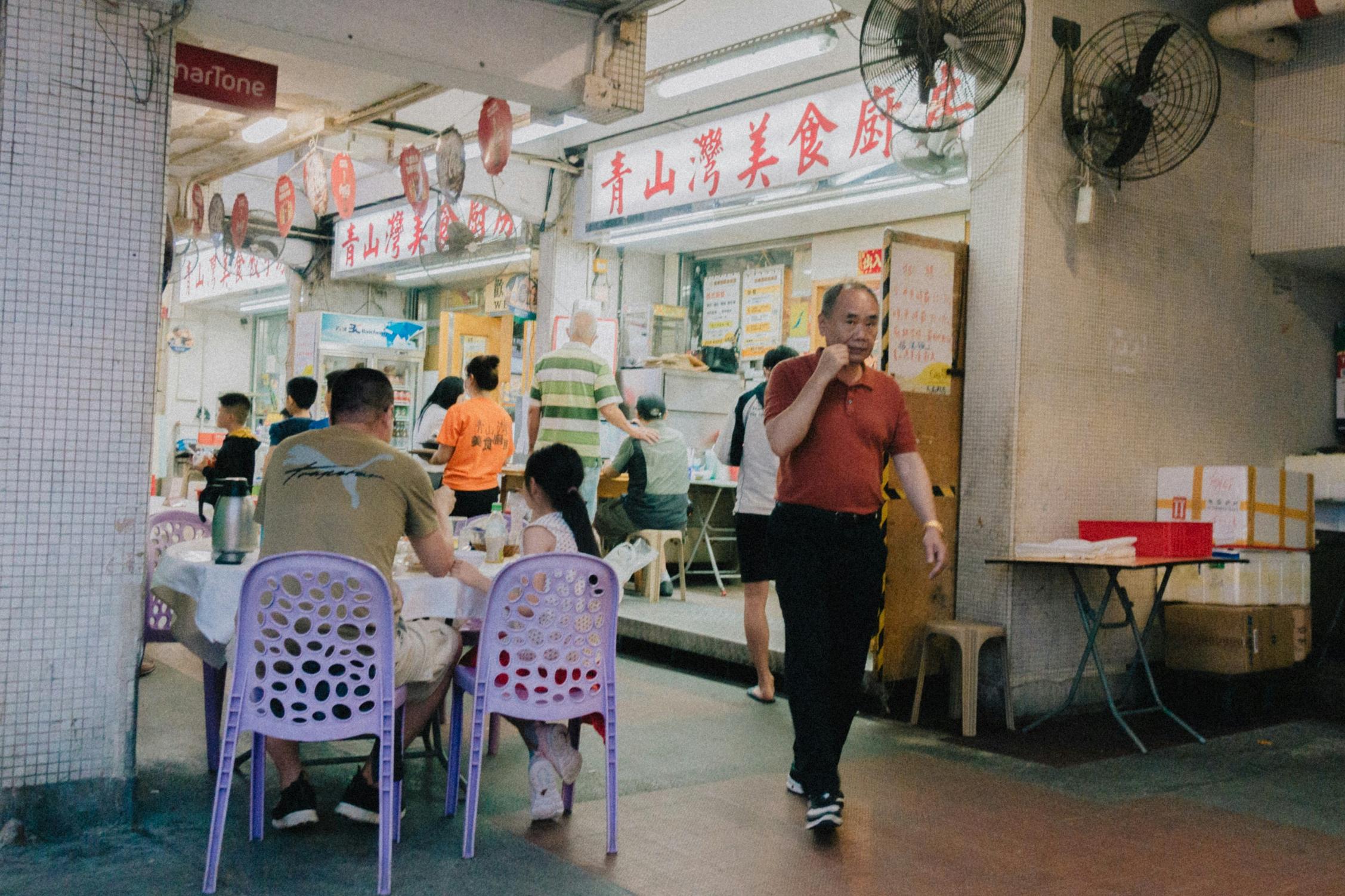 This candid scene captures a casual street-side eatery where diners sit on purple plastic chairs at round tables to enjoy their meals. A man in a red polo shirt walks past the bustling space, which is characterized by tiled walls, industrial fans, and prominent red Chinese signage hanging above the entrance.