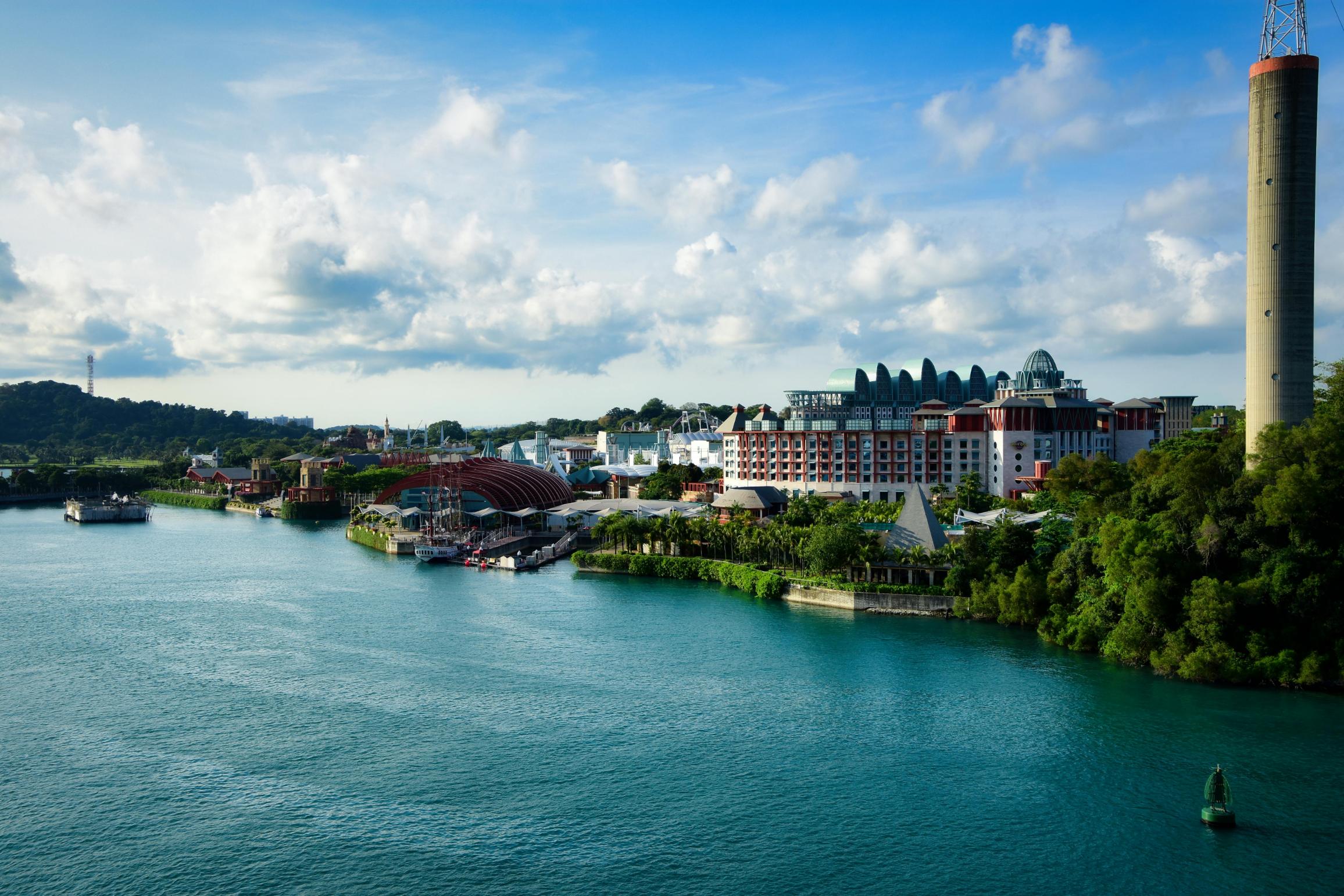 This scenic landscape captures a wide expanse of turquoise water bordering a lush coastline dotted with colorful resort buildings and unique architectural structures. A tall, slender concrete tower anchors the right side of the frame, rising towards a bright blue sky filled with scattered white clouds.