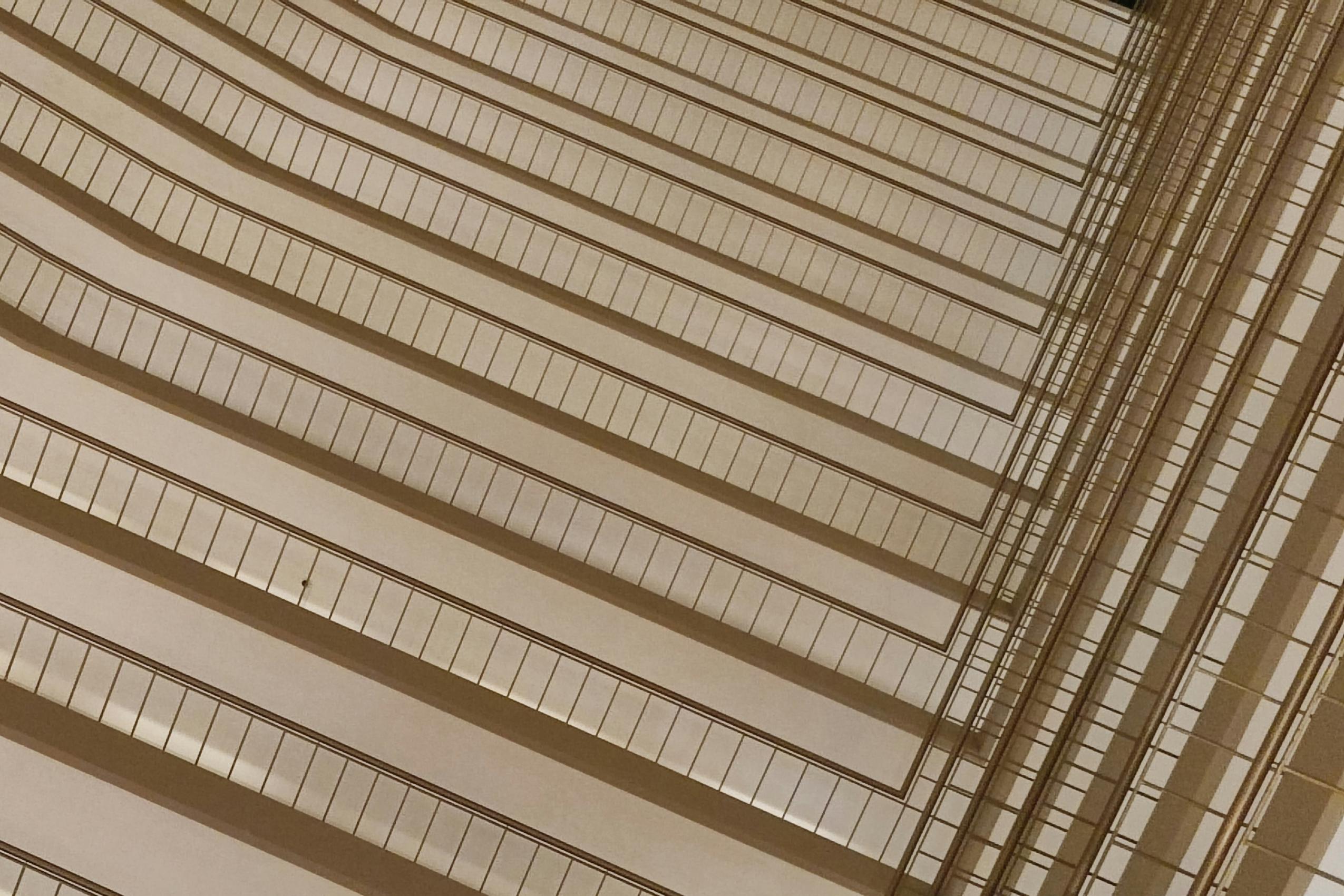 This abstract architectural composition captures a repetitive series of building levels viewed from below, creating a mesmerizing pattern of parallel diagonal lines. The warm, monochromatic beige tones highlight the geometric precision of the railings as they converge toward a sharp structural corner.