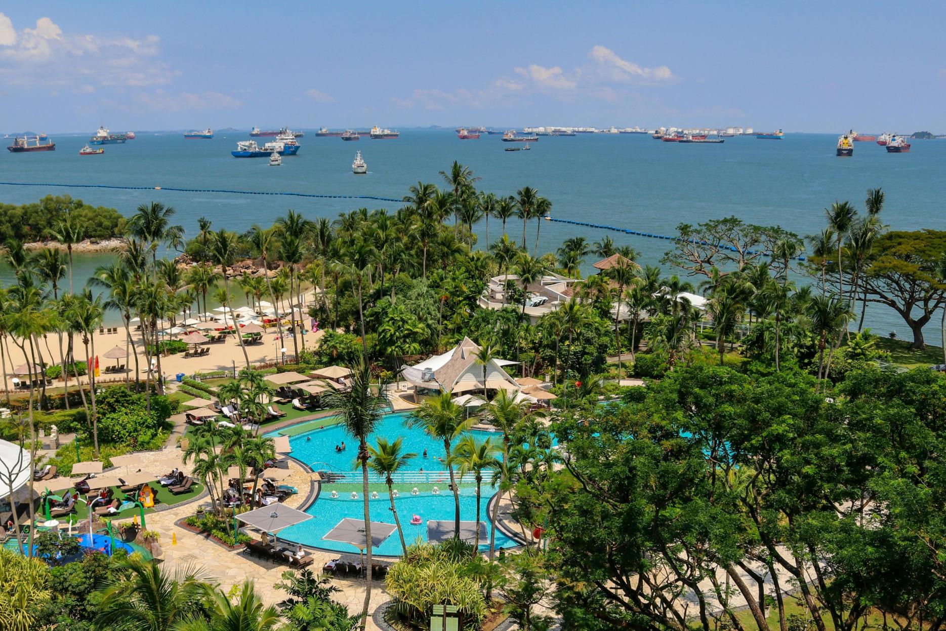 This aerial view depicts a vibrant resort complex featuring a large, blue swimming pool and sandy beach area surrounded by lush tropical greenery. In the distance, the calm ocean waters are heavily populated with a multitude of cargo ships and tankers stretched across the horizon under a sunny sky.