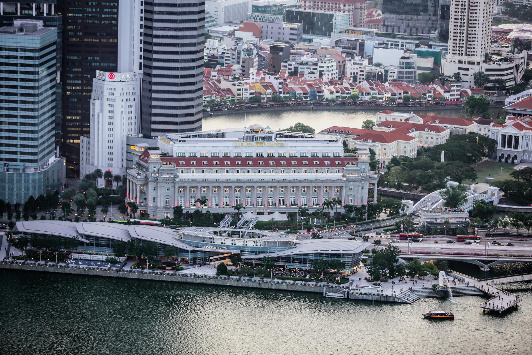 This high-angle urban view centers on the historic Fullerton Hotel, featuring a grand neoclassical facade and red roof, juxtaposed against towering modern skyscrapers on the left. The scene extends to a winding river lined with lower red-roofed buildings in the background and a busy waterfront promenade with a contemporary low-rise complex in the foreground.