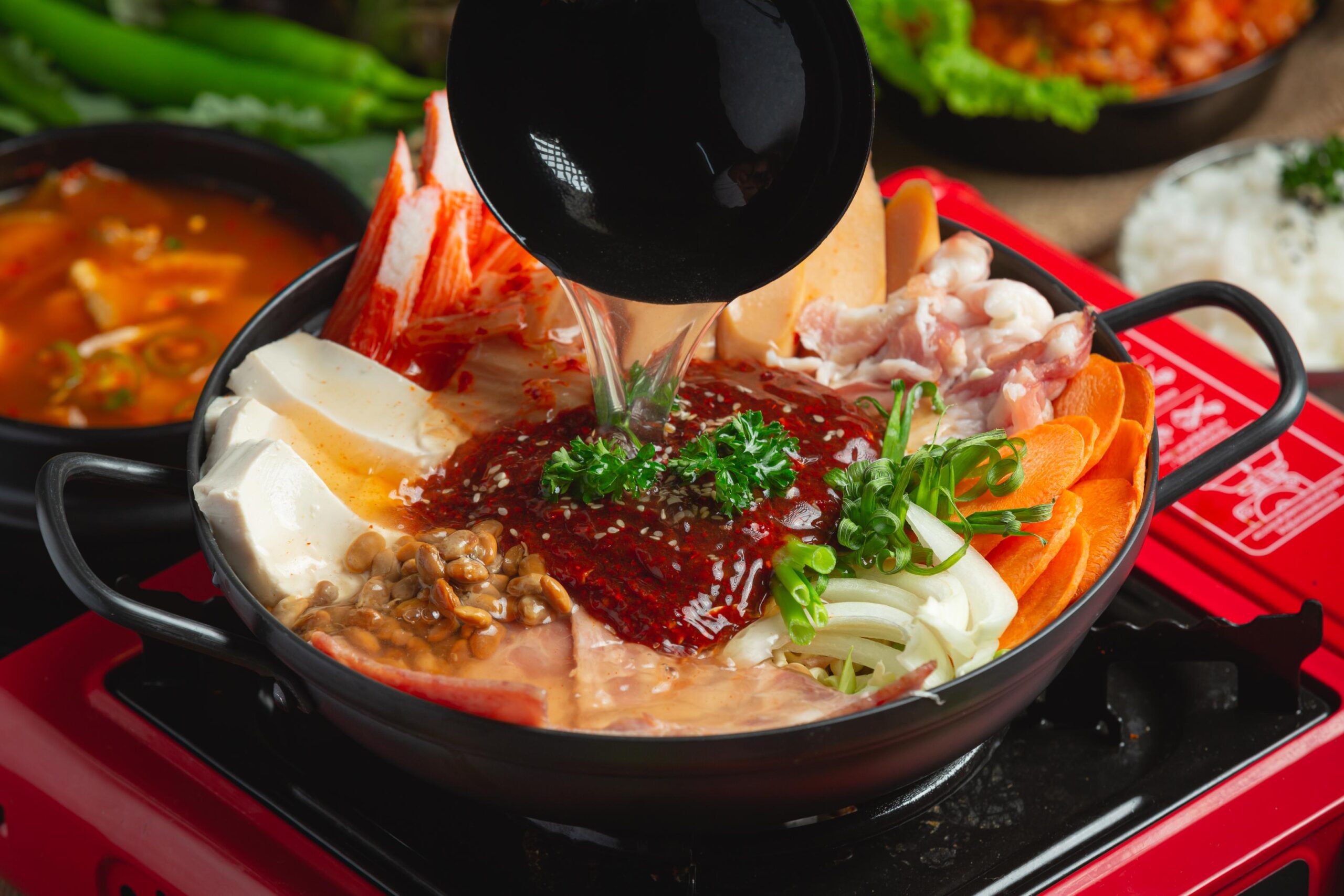 Clear broth is being poured from a ladle into a black pot filled with an assortment of ingredients, including tofu, sausages, ham, crab sticks, baked beans, and vegetables arranged around a dollop of spicy red paste.