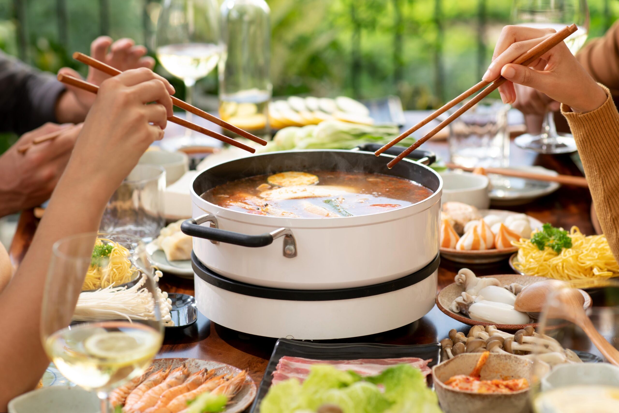 A group of friends shares a communal meal, using wooden chopsticks to reach into a simmering white hot pot filled with savory red broth. The table is lavishly spread with plates of fresh ingredients like shrimp, mushrooms, sliced meat, and noodles, accompanied by glasses of white wine in a bright, airy setting.