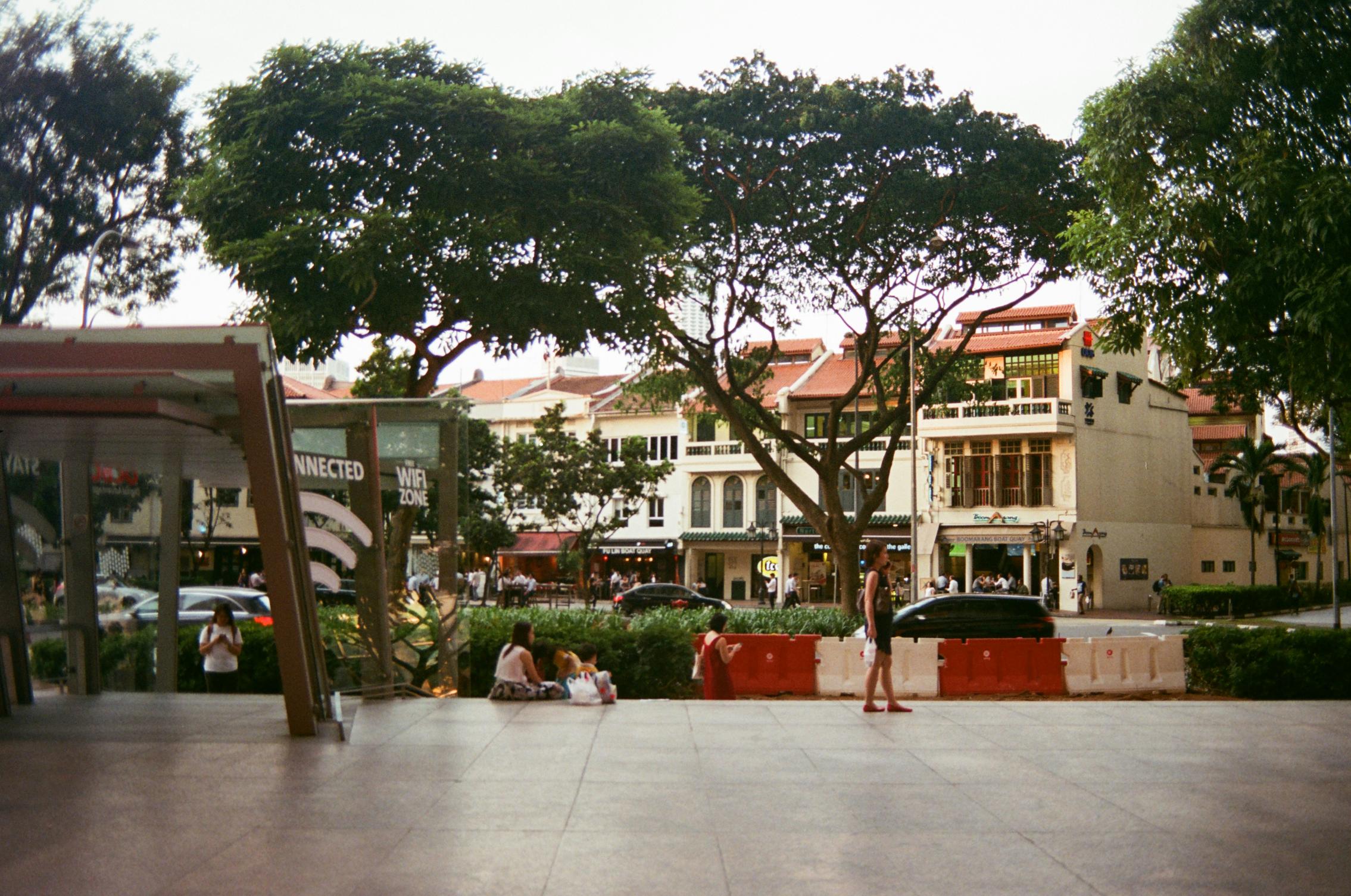 Pedestrians relax and walk near a modern glass shelter marked "WIFI ZONE" in the foreground of this bustling urban street scene. Behind a line of red and white road barriers, large sprawling trees shade a row of traditional white shophouses with red-tiled roofs.
