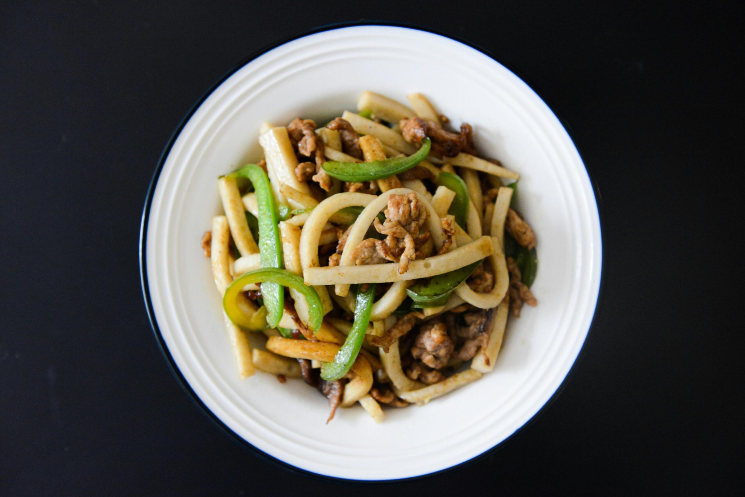 A top-down view presents a white bowl filled with a savory stir-fry consisting of thick noodles, sliced green bell peppers, and browned pieces of meat. The clean, ridged rim of the bowl stands out against the solid black background, highlighting the textures and colors of the dish.