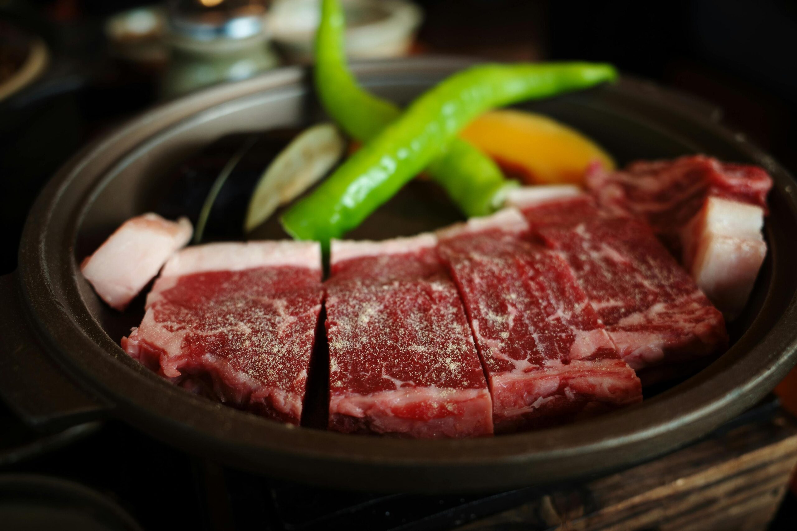 Thick slices of raw, marbled beef dusted with seasoning sit in a dark ceramic dish accompanied by fresh green peppers and vegetable slices like eggplant and pumpkin. The vivid red of the meat contrasts sharply with the bright green garnish, suggesting a high-quality meal ready for grilling or cooking.