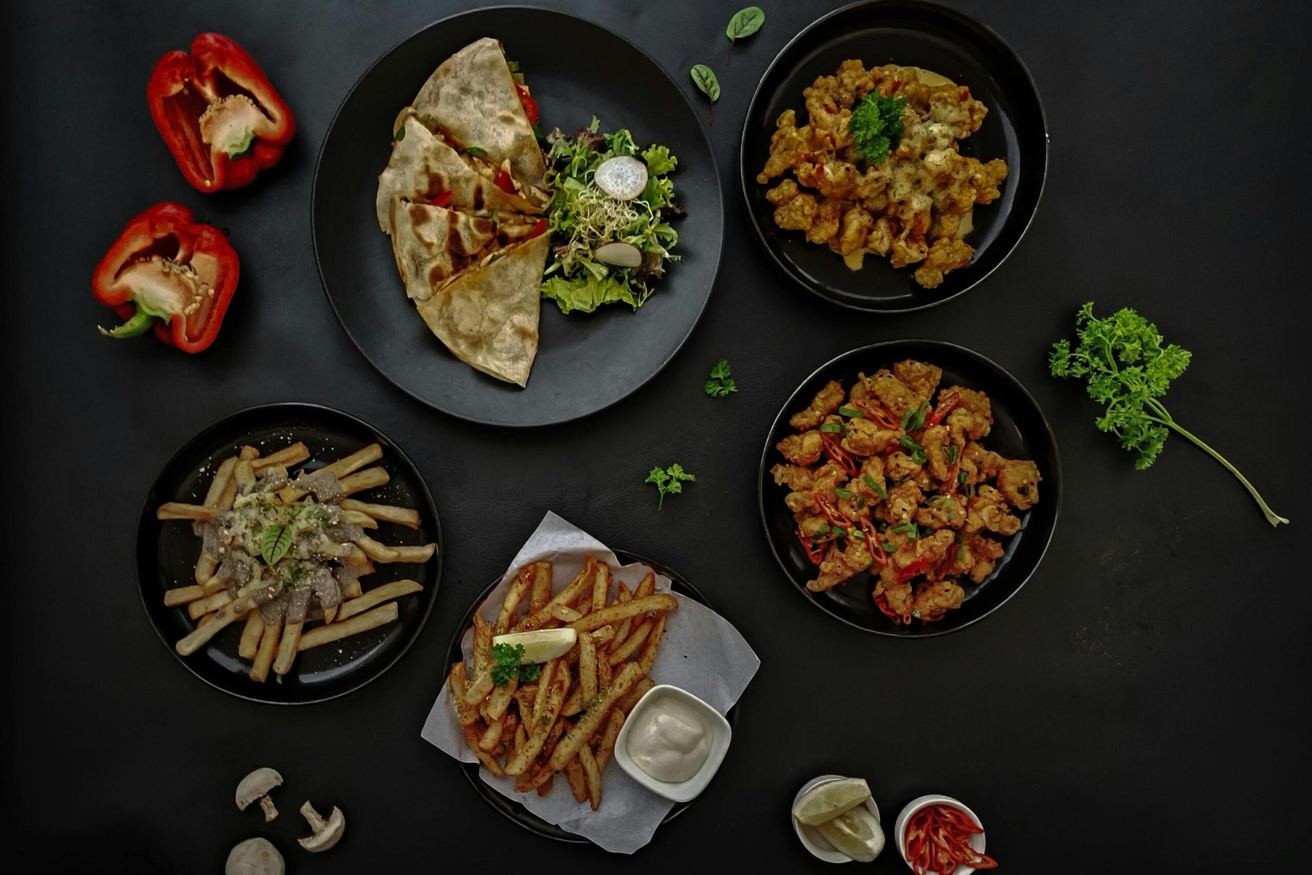 An assortment of savory dishes, including quesadillas, loaded fries, and two styles of fried chicken, is arranged on black plates against a dark background. Fresh garnishes such as red bell pepper halves, mushrooms, lime wedges, and parsley are scattered around the food to complete the flat-lay composition.