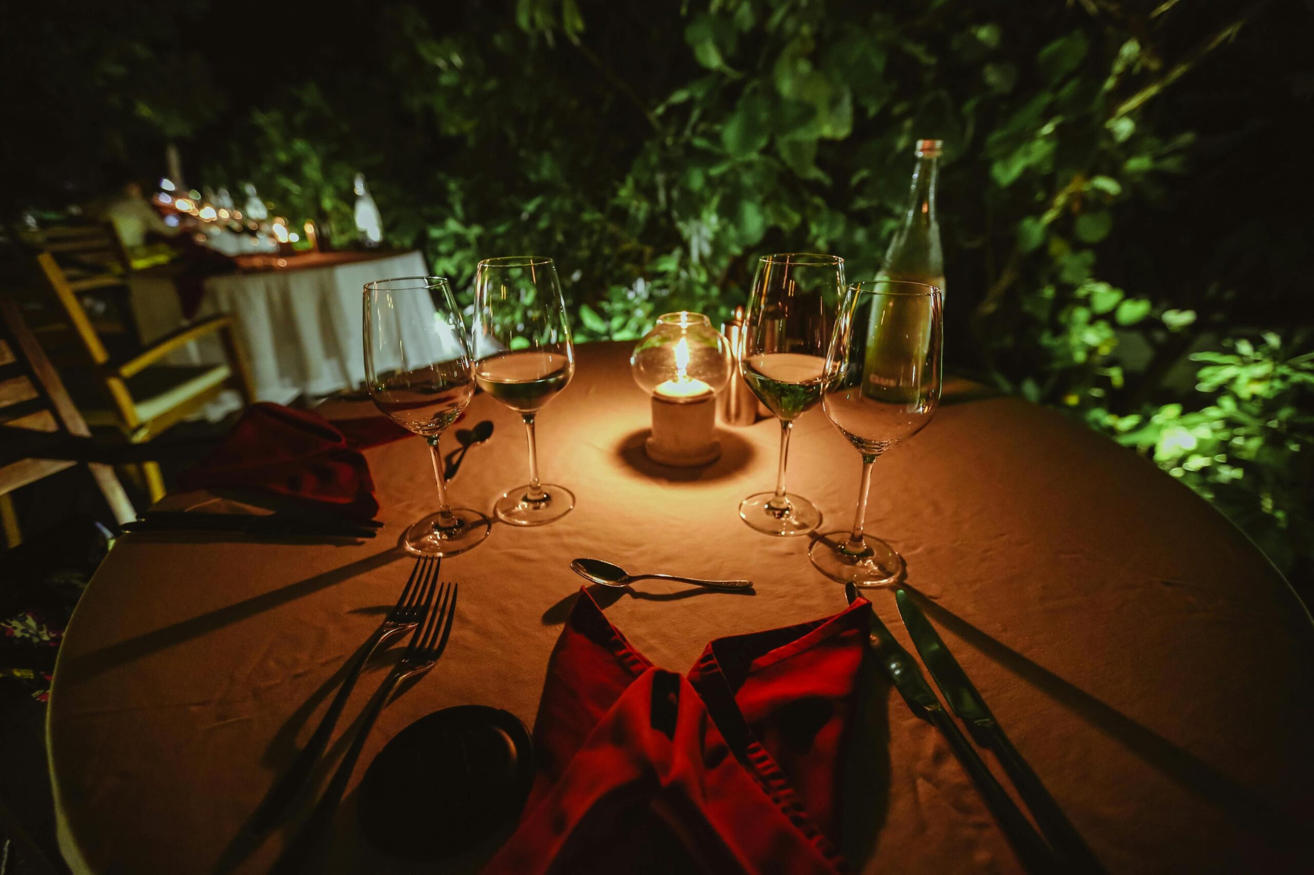 An intimate outdoor dining table is elegantly set for the evening with wine glasses, silverware, and red napkins, all bathed in the warm glow of a central candle. Lush greenery surrounds the scene in the darkness, creating a cozy and romantic atmosphere perfect for a dinner.