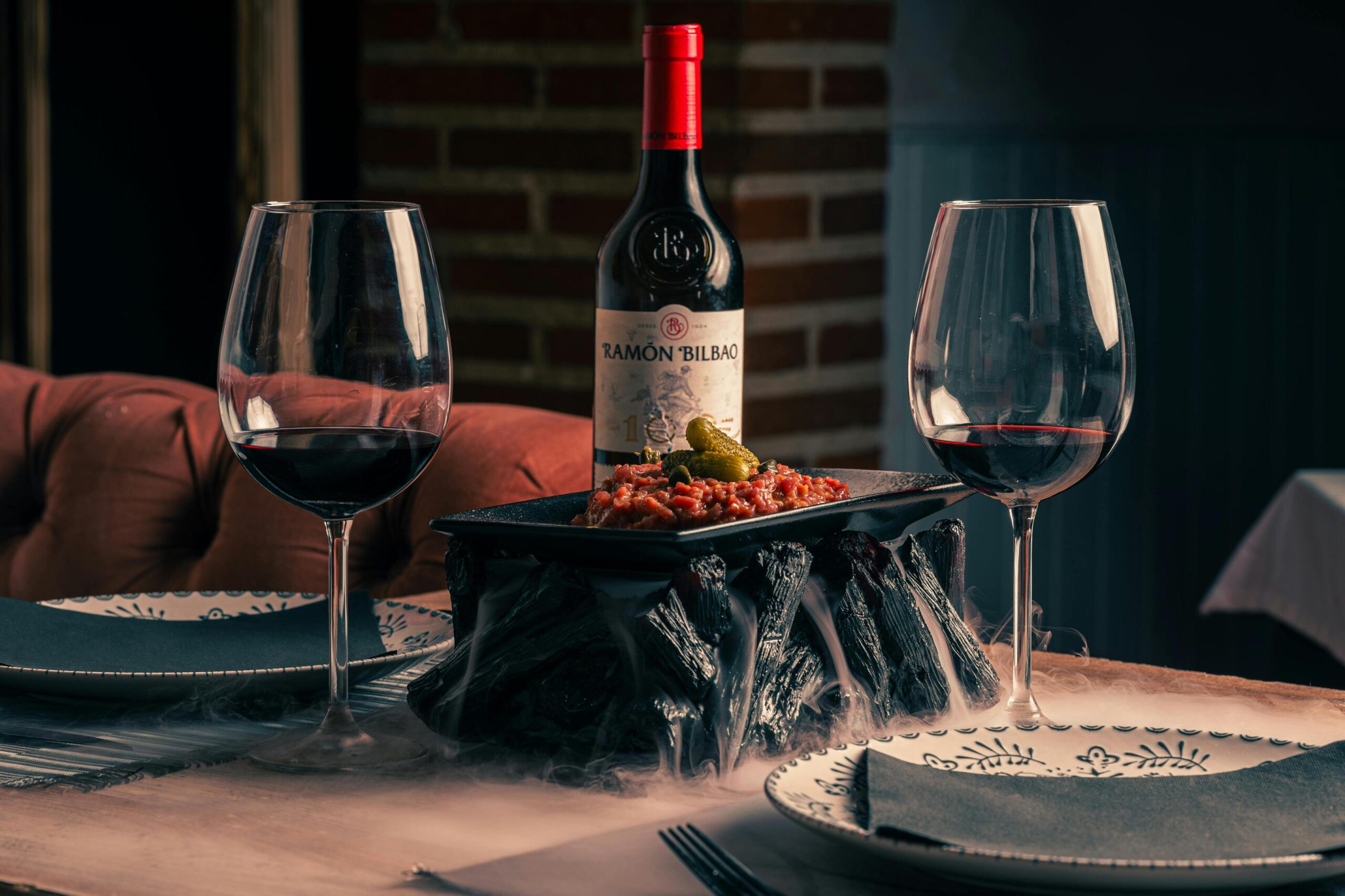 Two glasses of red wine flank a dramatic culinary presentation featuring a tartare-style dish served atop a bed of smoking charcoal-like pieces. A bottle of Ramón Bilbao wine stands centrally behind the food, contributing to the sophisticated and atmospheric dining setting.