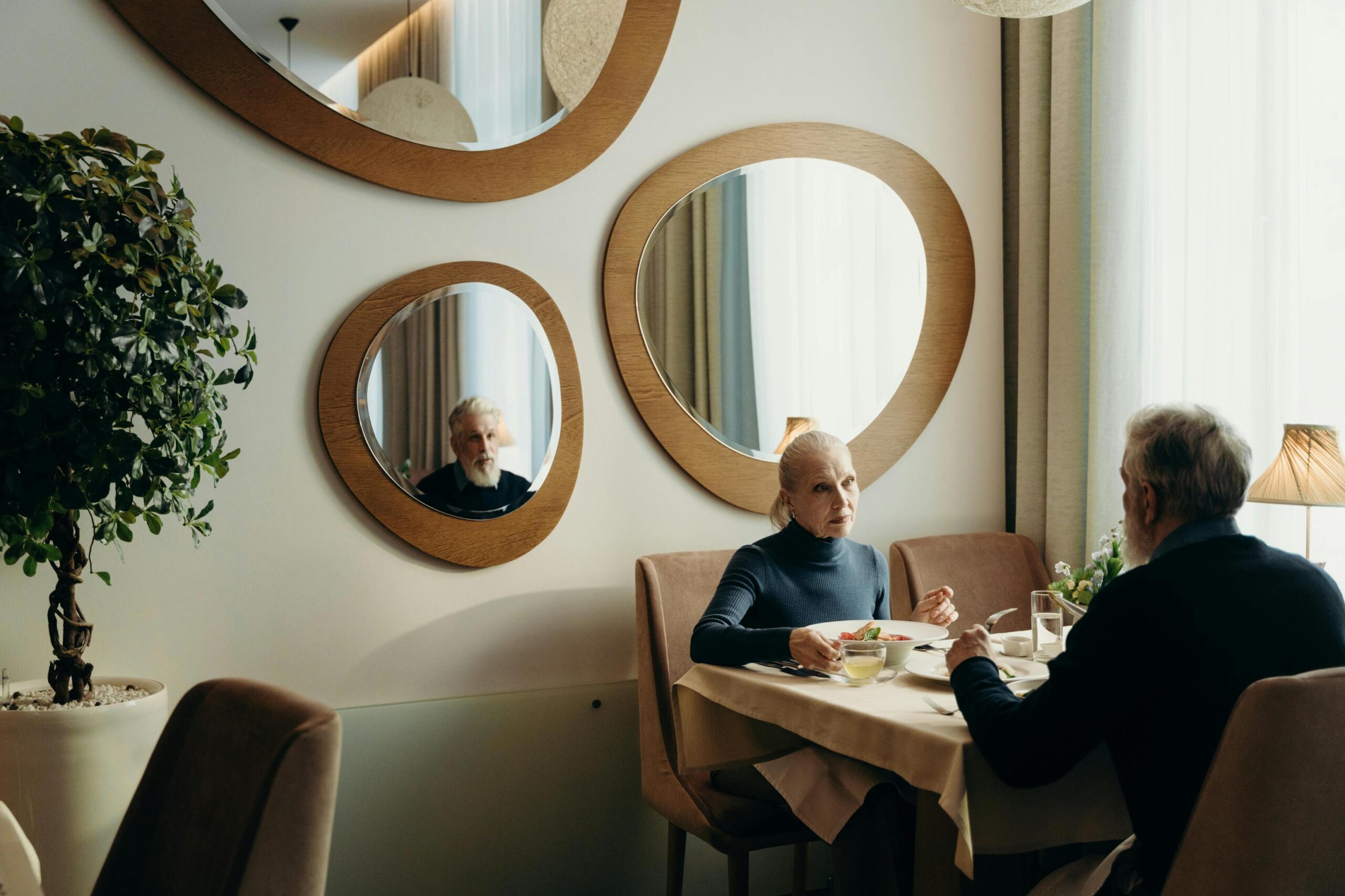 An elderly couple shares a quiet meal in a bright, modern dining room featuring unique, organic-shaped mirrors on the wall. The woman looks towards her partner, whose face is revealed to the viewer through his reflection in one of the mirrors, adding depth to the scene.