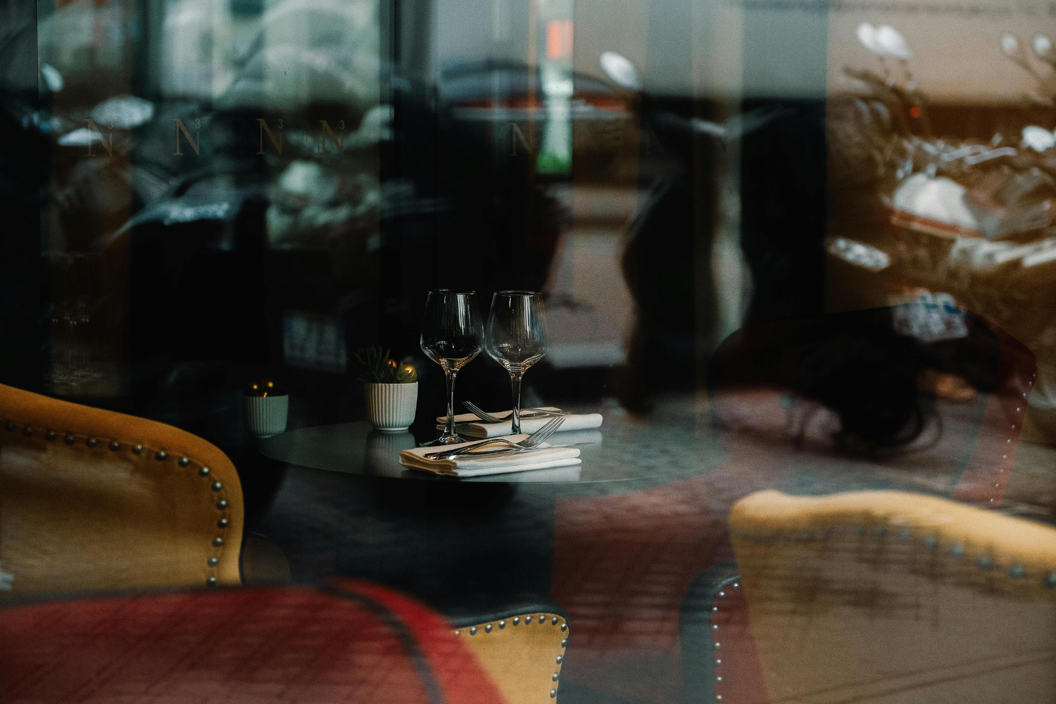 Through a reflective window pane, a small round table is neatly set with wine glasses and silverware, creating a quiet moment amidst the ghostly reflections of the street. Plush upholstered chairs in mustard and red tones frame the foreground, emphasizing the cozy and inviting atmosphere of the restaurant interior.