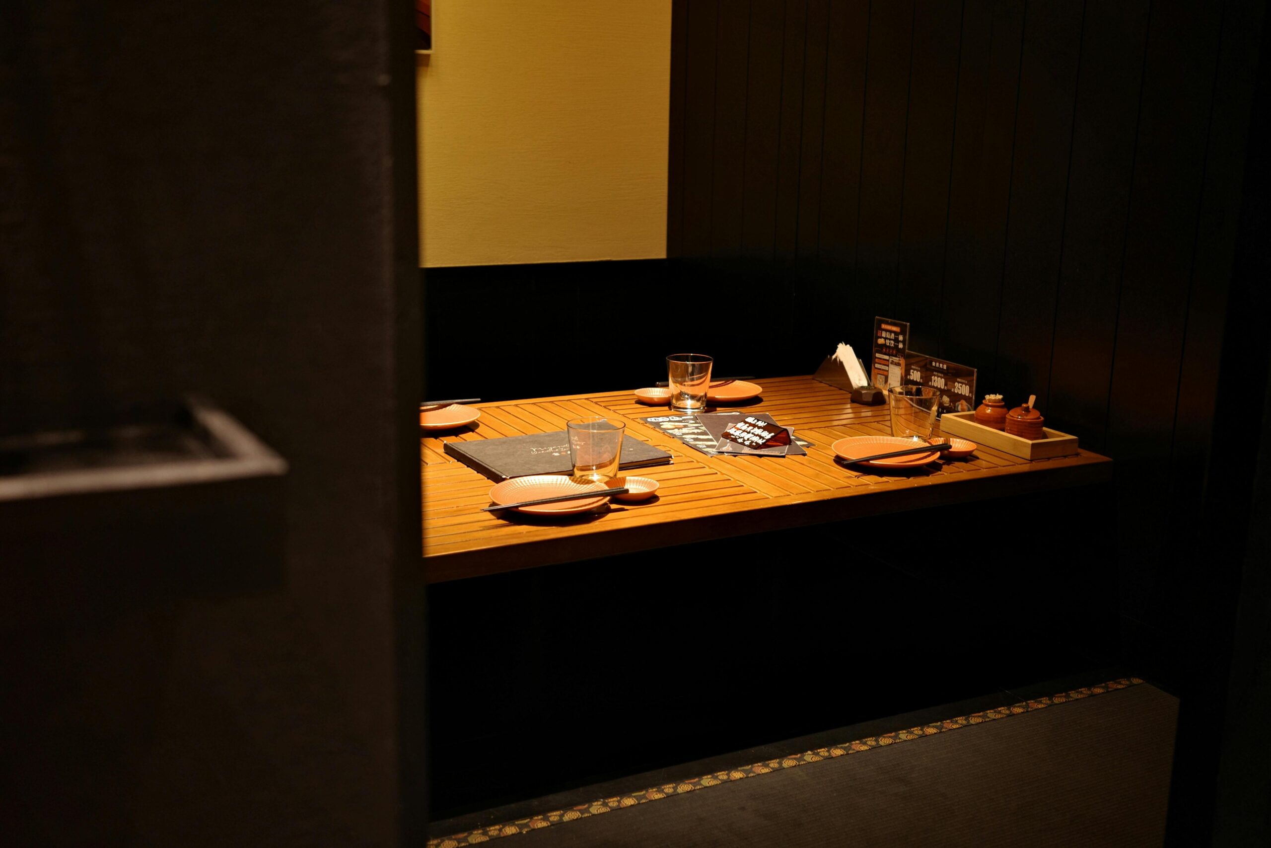 A wooden table is bathed in warm light within a dark, private restaurant booth, highlighting the clean setup of terracotta plates and glassware. Chopsticks, menus, and condiment jars are neatly arranged on the slatted surface, inviting diners into the quiet and moody space.