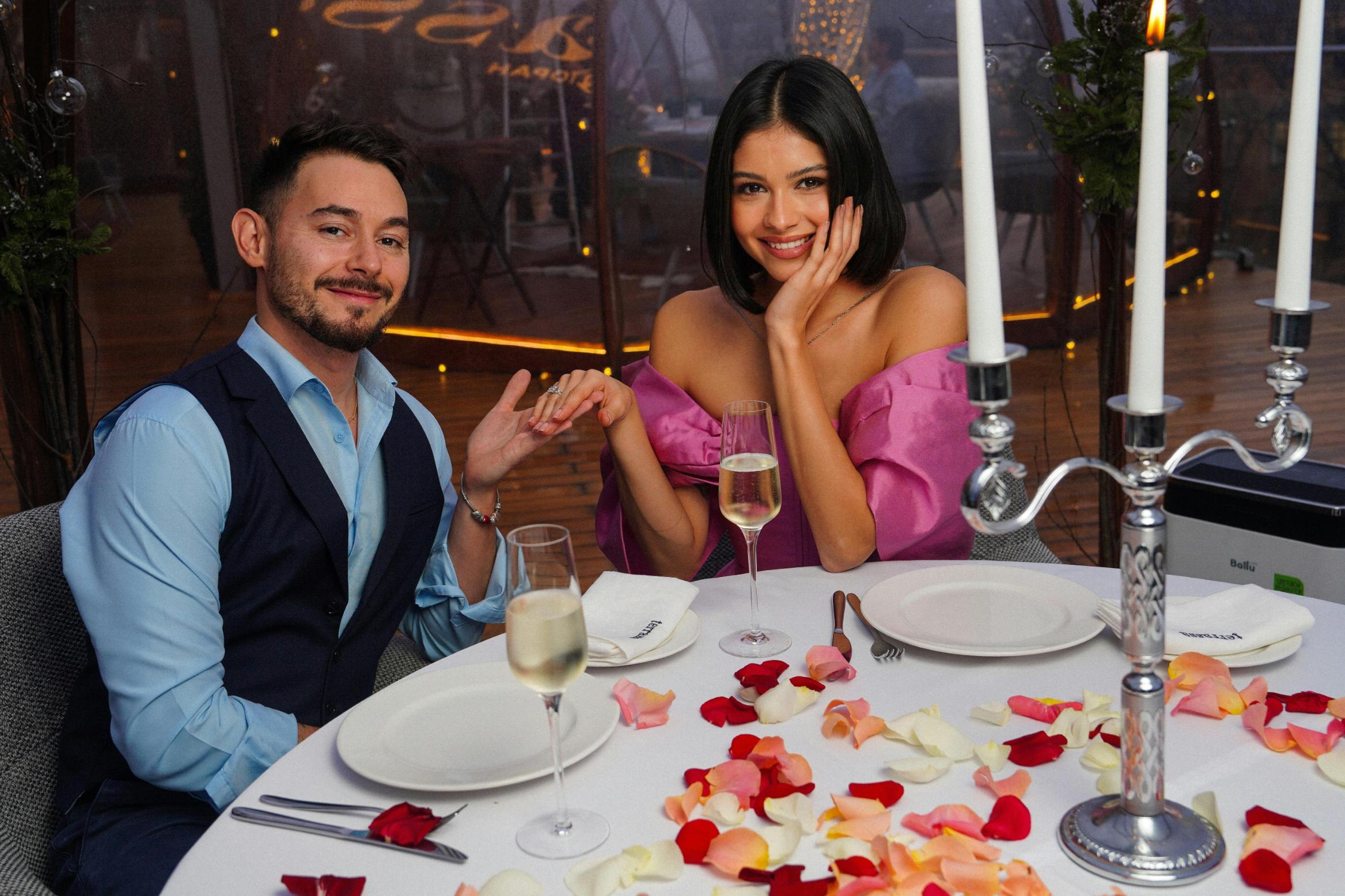 A smiling couple celebrates a romantic occasion at a table beautifully strewn with multi-colored rose petals and set with a tall silver candelabra. The man gestures towards the woman's hand, highlighting an engagement ring, as they pose before glasses of champagne in a festive evening setting.