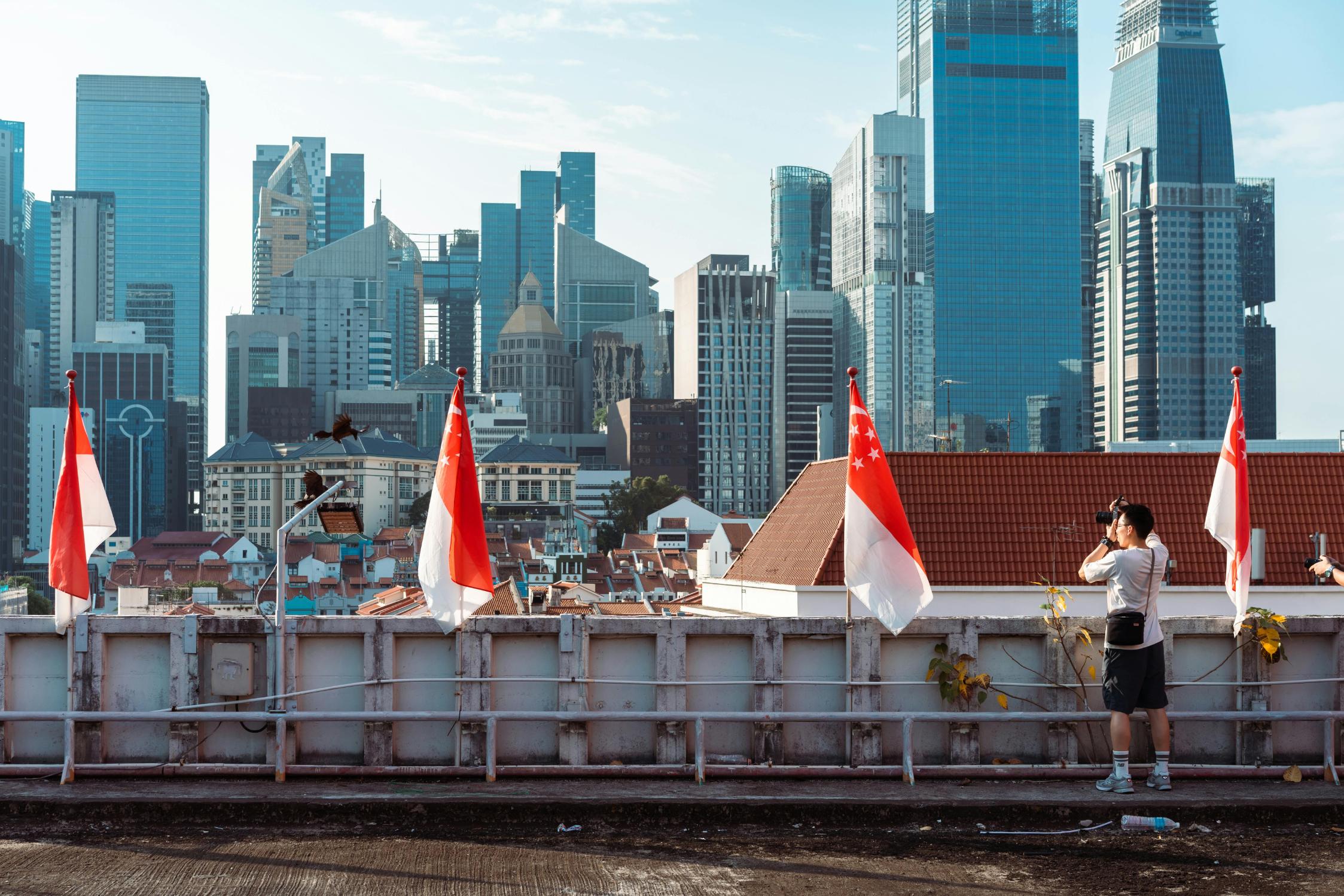 A photographer stands on a rooftop lined with Singapore flags, capturing the vibrant city skyline under a clear blue sky. The view highlights a striking architectural contrast, featuring traditional red-tiled roofs in the foreground set against a backdrop of towering modern skyscrapers.