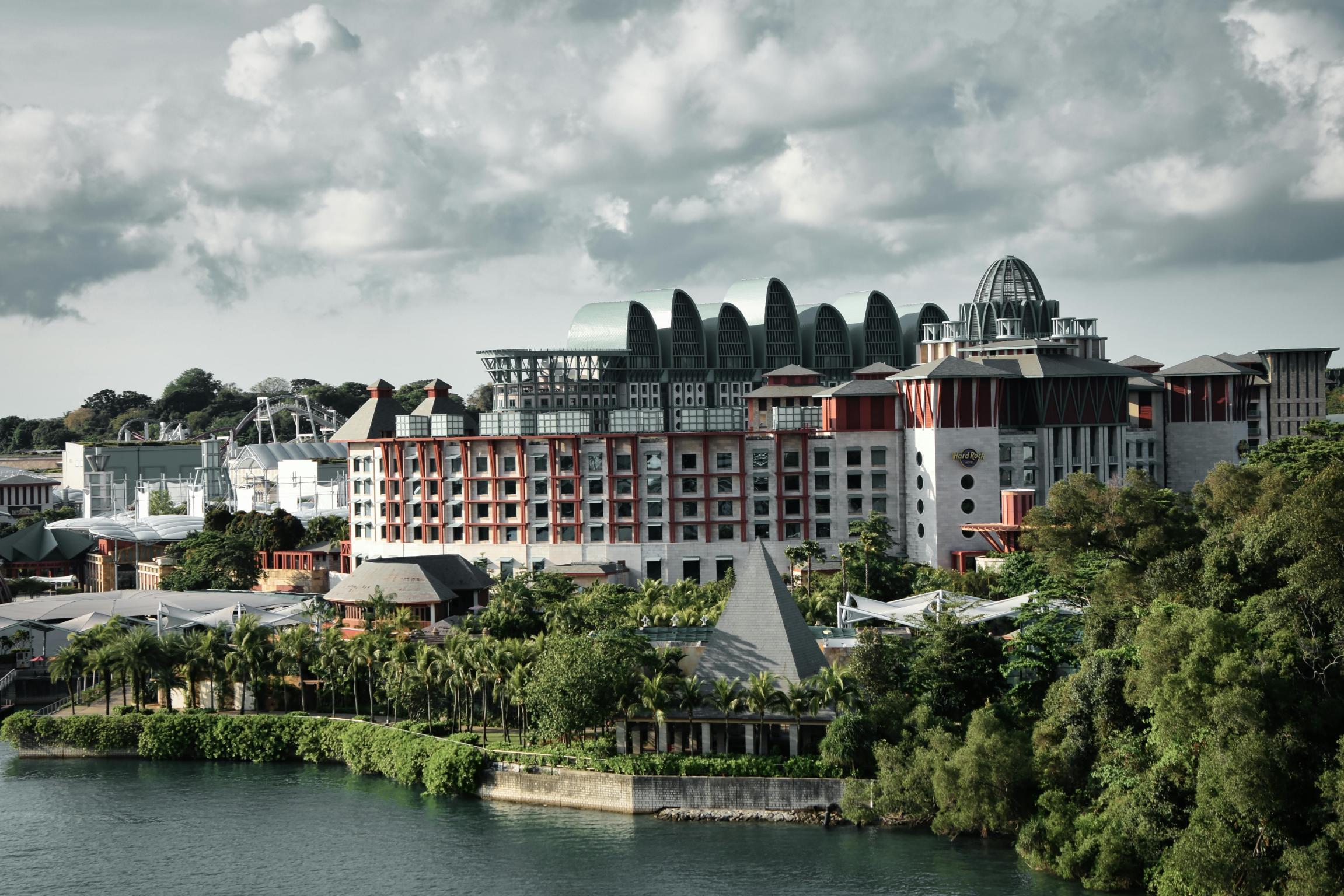This image features a sprawling resort complex with distinctive wave-like roof structures and red architectural accents, nestled amidst lush tropical greenery along a waterfront. Behind the modern buildings and palm trees, roller coaster tracks are visible in the distance against a backdrop of dramatic, cloudy skies.