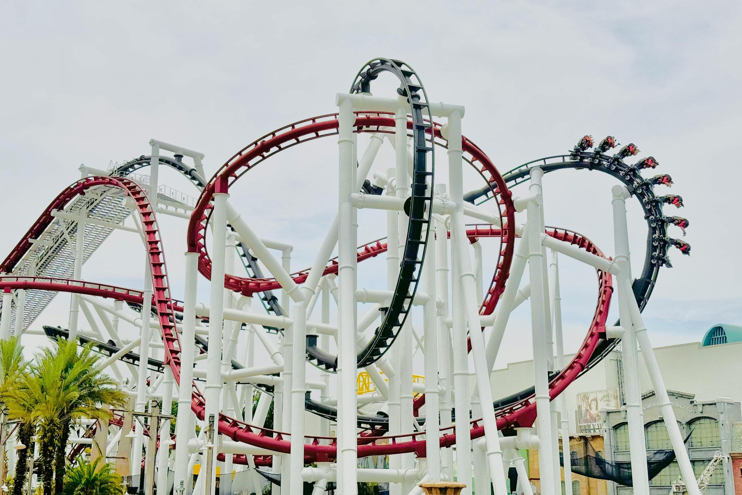 This image features a complex roller coaster with twisting red and black tracks weaving through a massive white support structure. A train carrying passengers is captured navigating a high-speed loop, set against a cloudy sky with palm trees visible near the base.